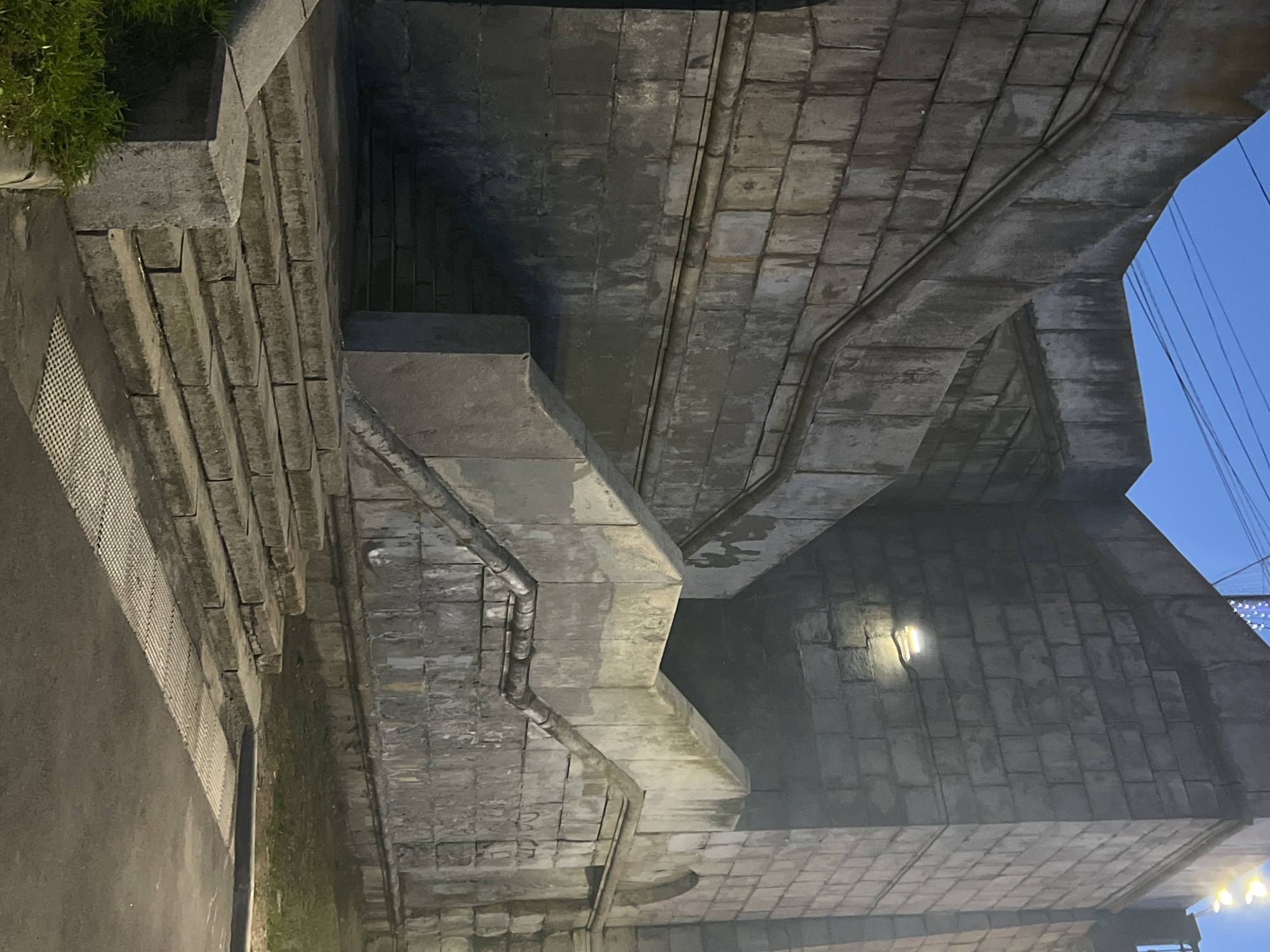 The image depicts a stone staircase leading up to a weathered, brick-like structure, likely part of a bridge or embankment, in Tver, Russia. The sky above is a deep twilight blue, with visible power lines stretching across the frame. Several lights are illuminating parts of the structure, including a bright, modern-looking lamp mounted on the wall. The stone steps are worn and cracked, with patches of grass growing at the base. A textured metal grate is visible on the paved area in front of the steps. The overall impression is one of an aged, utilitarian urban landscape, possibly a pedestrian walkway or access point. There are no people or animals visible in the scene.