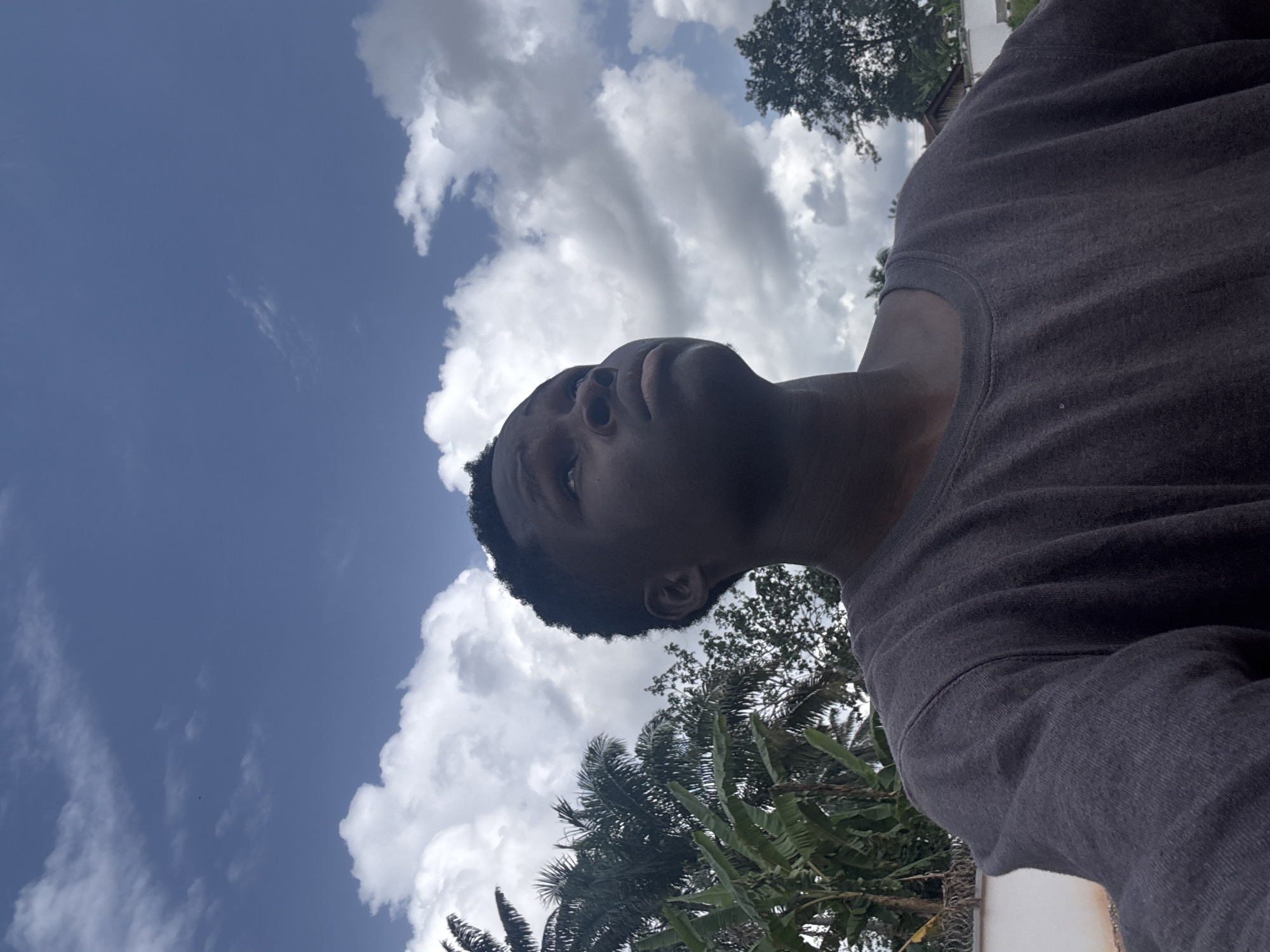The image is a low-angle selfie of a person with dark skin and short hair wearing a gray long-sleeved shirt. The person is looking upwards. Behind the person, there is a sky with white clouds and blue sky. There are trees, including palm trees, visible. The location is likely outdoors in a tropical environment; however, it's impossible to confirm if the location is actually Eziudo, Nigeria, based on the image alone. There are no other people or objects visible in the frame. The action is the person taking a selfie looking up. The interaction is between the person and their surroundings.