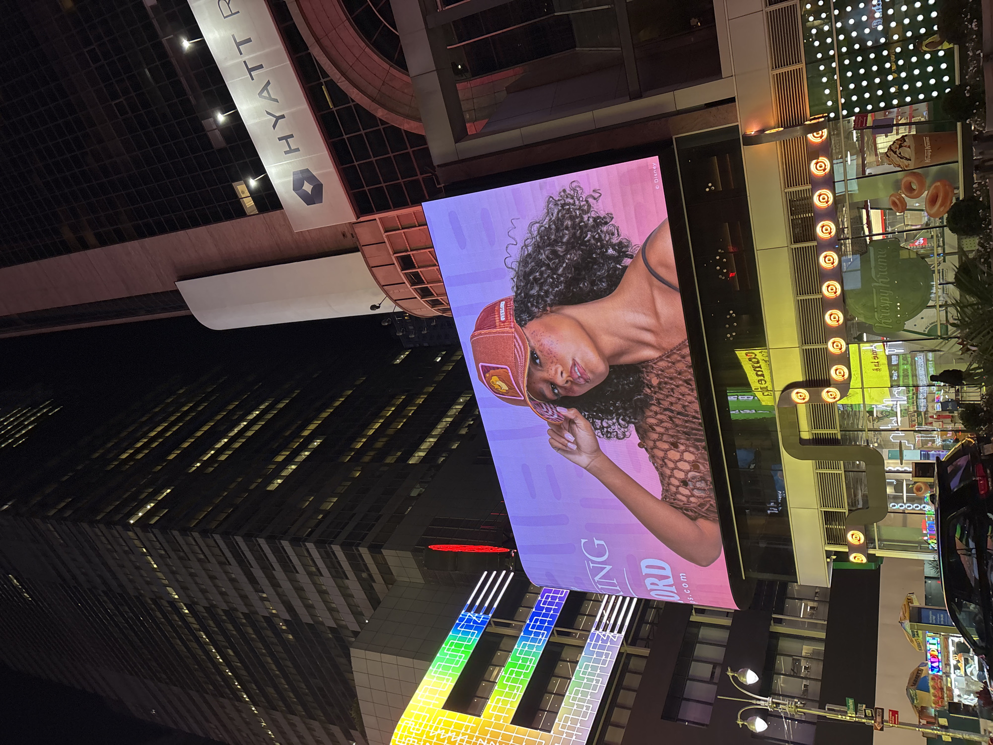 This media file captures a vibrant, high-density urban street scene at night, characterized by extensive artificial lighting and large digital advertisements. The perspective is tilted, but when viewed upright, it showcases a bustling commercial area.
The primary subject is a massive digital billboard occupying the central-right portion of the frame. It displays an advertisement featuring a woman with curly dark hair, wearing a brown cap and a textured top, looking towards the viewer. The background of the billboard is a soft gradient of purple and pink. Visible text on this screen includes "ING ORD s.com", "OVERLORD", and a small copyright notice "© Disney". Directly below this billboard is a brightly lit "Krispy Kreme" storefront, showcasing donuts and beverages in its windows, with its distinctive green logo prominent.
To the upper left, a tall building features an illuminated sign for "HYATT R" (likely Hyatt Regency) with its corporate logo. The surrounding architecture includes several dark skyscrapers, some with visible illuminated windows, contributing to the metropolitan backdrop. In the lower left, an elaborate, multi-tiered geometric structure glows with a vibrant rainbow of colors, adding another layer of light and visual interest to the scene. A standard two-lamp streetlight is also visible, typical of an urban environment.
The overall impression is one of an active, commercial hub after dark, likely a tourist or entertainment district, given the scale and number of advertisements and brand presences. The scene is devoid of specific events, instead offering a snapshot of a lively nocturnal city street.
