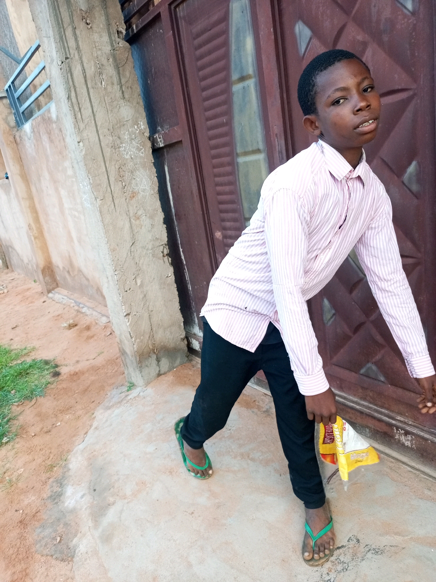 A young boy, appearing to be of Nigerian descent and approximately 10-12 years old, stands outdoors near a brown metal door. He is wearing a pink and white striped long-sleeved collared shirt tucked into black pants. His feet are clad in green flip-flops. In his right hand, he holds a yellow and white plastic bag. The ground is a mix of dirt and concrete. To the left of the door, a section of a textured, light-colored building wall is visible, featuring some horizontal metal bars above. The scene appears to be in Tempure, Nigeria.