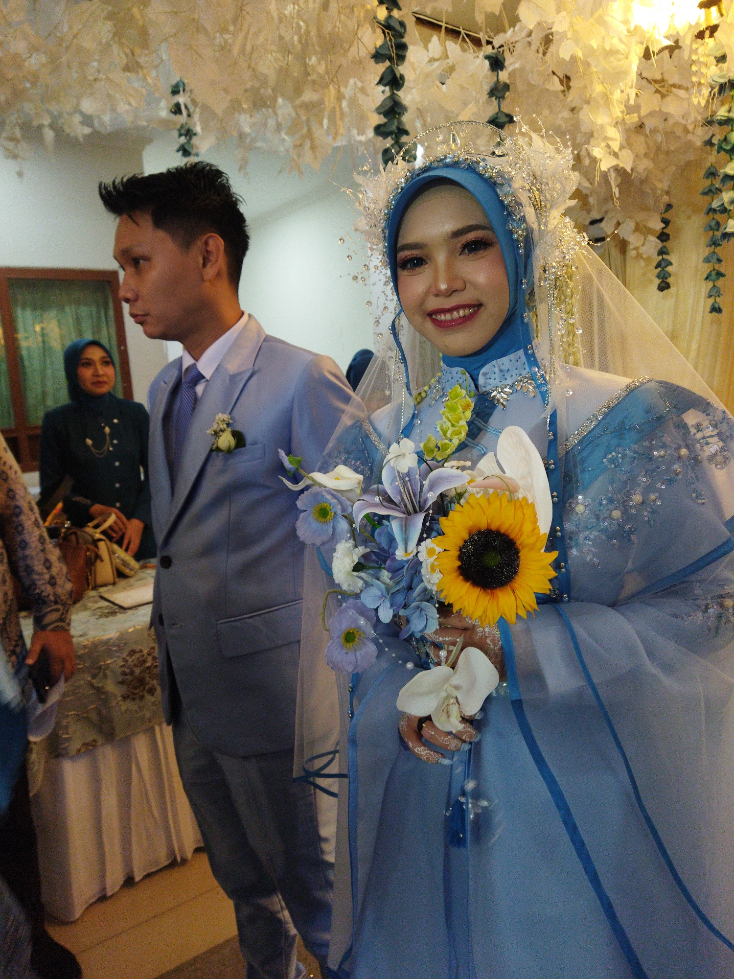 A formal indoor wedding scene is depicted, likely in Semarang, Indonesia, given the clothing and cultural context. A man in a light blue suit with a white shirt and blue tie stands to the left, looking to his left. His suit has a boutonnière of white flowers and greenery in the lapel. Beside him, to the right, is a woman dressed as a bride, wearing a blue hijab and a white and blue wedding gown adorned with intricate beading and embellishments. She is smiling and holding a bouquet that includes a large sunflower, lilies, and other blue and white flowers. Behind the groom, a woman in a dark teal hijab and matching outfit is visible, standing near a table covered with a patterned cloth. The environment is decorated with white floral arrangements and hanging greenery, suggesting a wedding ceremony or reception. The lighting is warm, indicating an indoor setting, possibly a banquet hall or a decorated room.