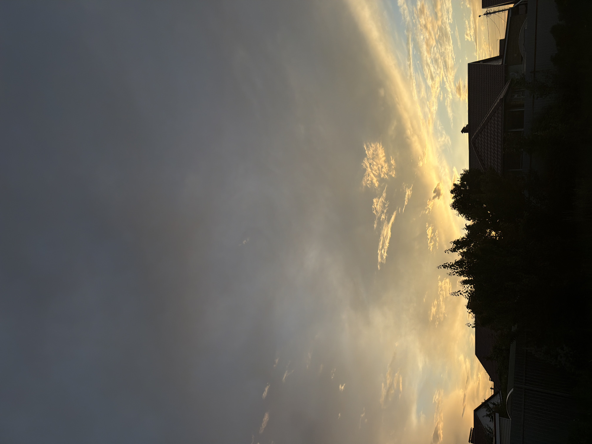 The image depicts a suburban or rural residential area at dusk, likely in Узюково, Russia, given the context. The sky is filled with clouds, transitioning from a deep grey at the top to a warm orange and yellow glow near the horizon, indicating either sunset or sunrise. Silhouetted against this illuminated sky are the rooftops of several houses and the dark foliage of trees. A fence runs across the foreground, obscuring the lower portions of the structures and vegetation. The scene is peaceful and tranquil, with no people or visible activity.