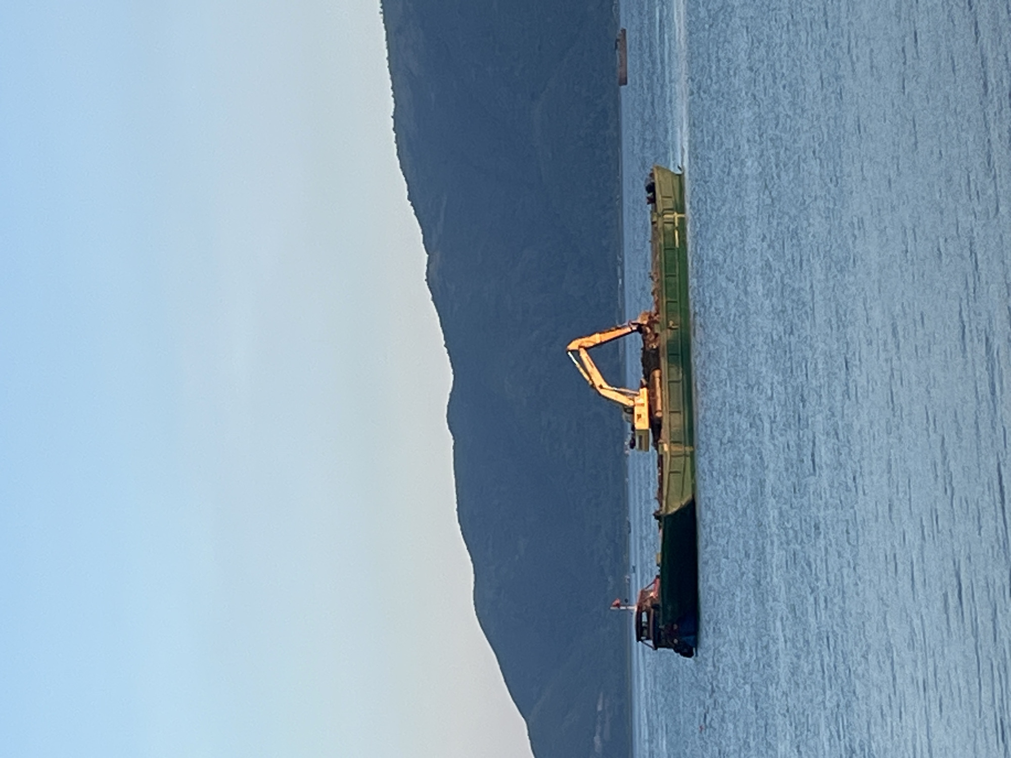 The image shows a dredging vessel in the water near the coast. The vessel is green and has an orange excavator arm on board. A mountain range is visible in the background. The sky is light blue. Given the location of Nam Nha Trang Ward, Vietnam, the water is likely a bay off the coast.