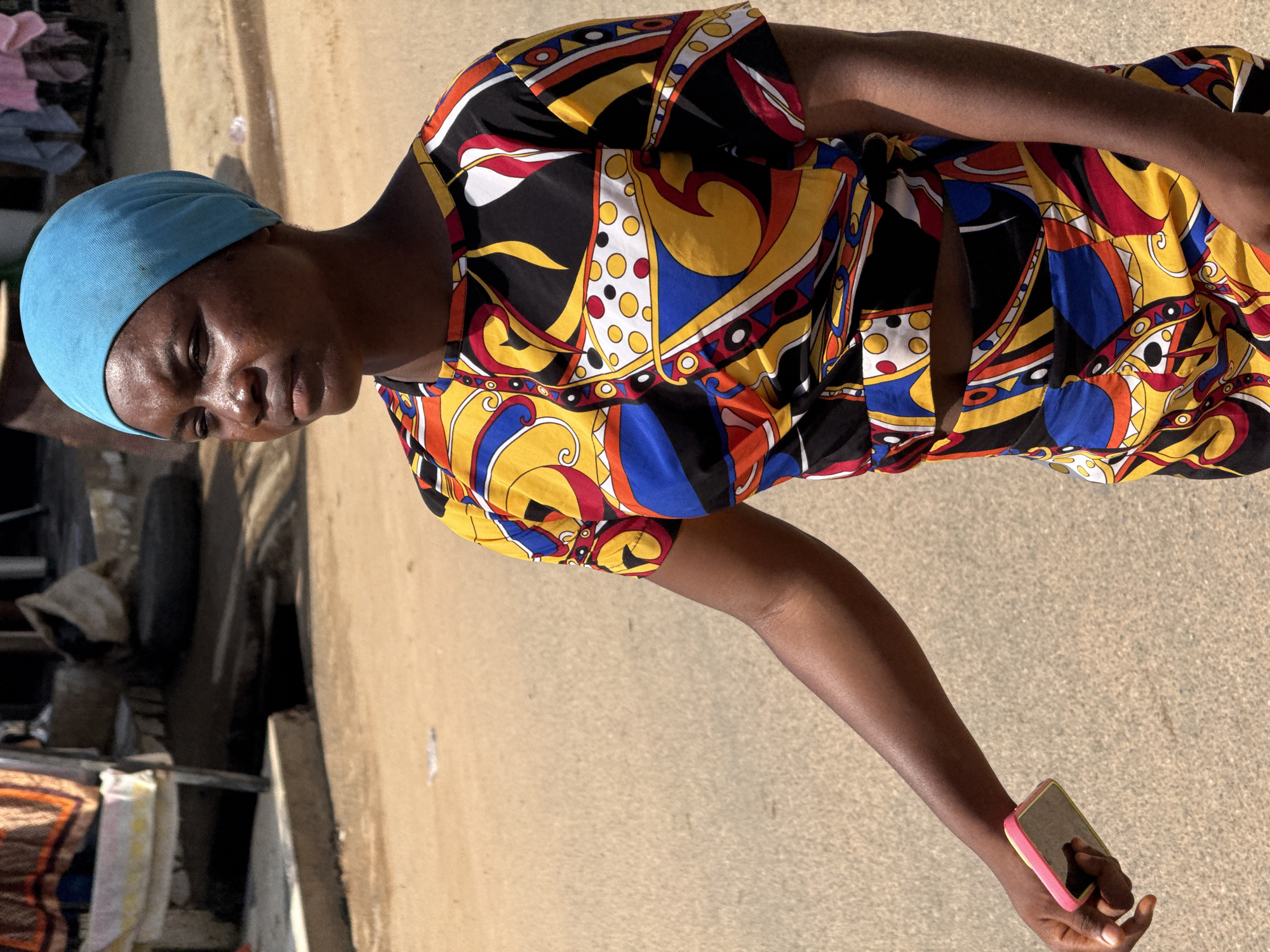 An outdoor medium shot shows a young Black woman with dark skin and brown eyes walking on a sandy road. She is wearing a bright, multi-colored short-sleeved shirt with a geometric pattern of yellow, blue, red, white, and black. She also wears a light blue headscarf. Her left hand is extended out, holding a pink smartphone. The background shows a dusty, unpaved road with some debris and structures to the left. The scene appears to be in a bustling urban environment. The image was taken in the city of Abuja, Nigeria.