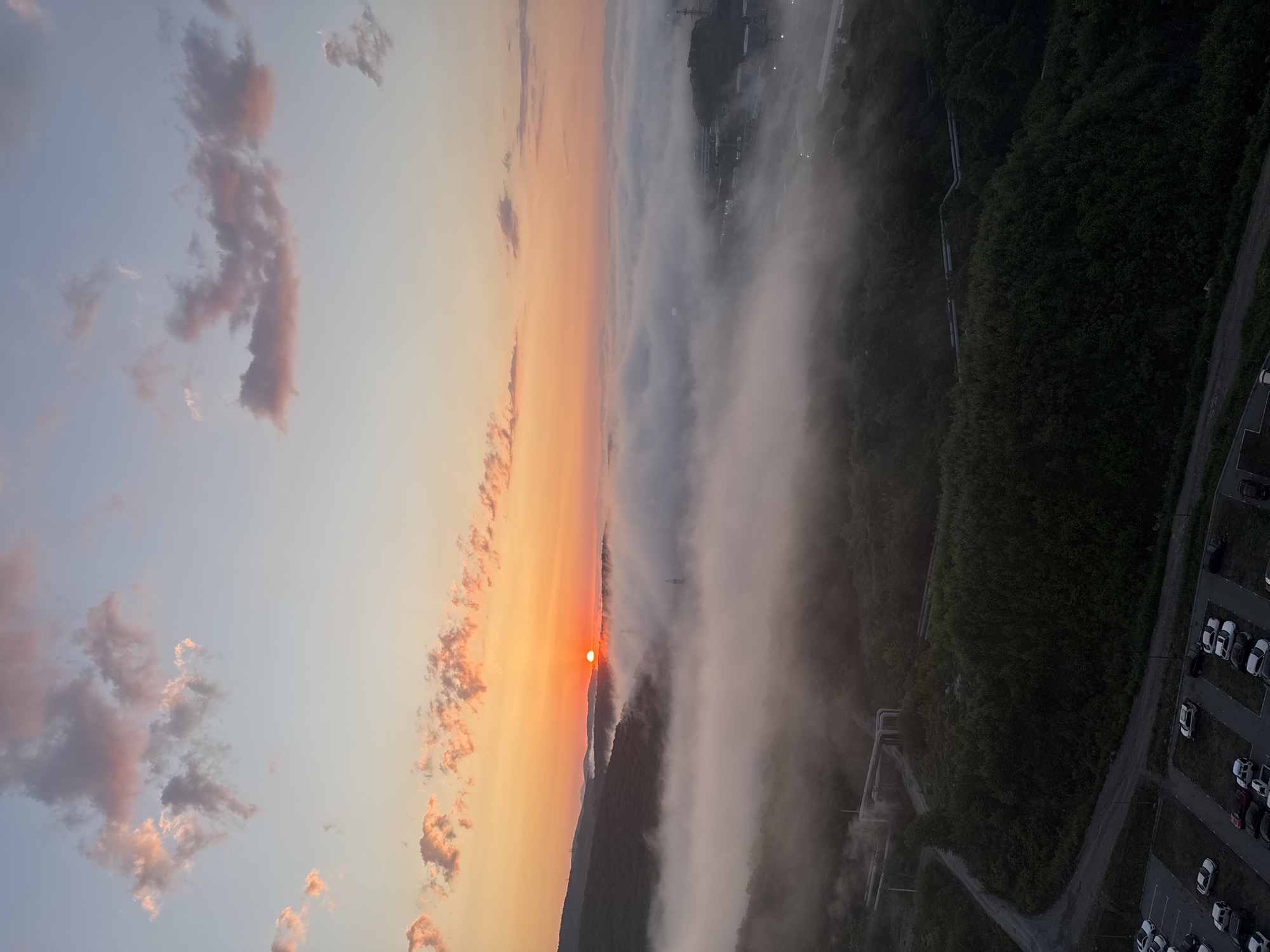 An aerial view captures a sunrise over Vladivostok, Russia, with a thick blanket of fog obscuring a valley. The sun, a bright orange orb, is positioned near the horizon, casting a warm glow across the sky and illuminating the edges of the clouds. The fog rolls in waves, partially covering forested hills and a distant industrial area with buildings and power lines. In the foreground, a parking lot is visible with several cars.