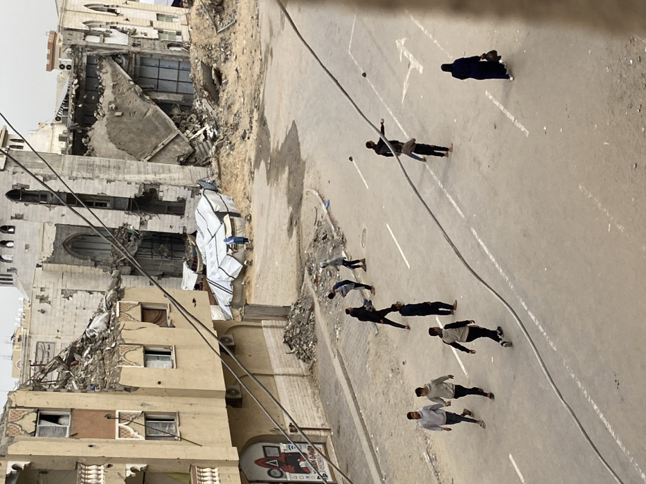 This image depicts a damaged urban street scene in Khan Yunis, Palestinian Territory. The foreground is dominated by a street with faded white lane markings and a dirt-covered shoulder on the left. Overhead, multiple thick utility cables traverse the frame diagonally.
On the left, a light brown multi-story building shows significant damage, including a collapsed upper section with exposed rebar and rubble. Its lower facade features decorative window frames, a white balcony railing, and what appears to be a storefront sign with red and black graphics resembling the "Squid Game" logo and text "TEEN GAMES".
Across the street, further into the background, two heavily damaged large stone buildings stand partially collapsed, with substantial portions of their structures missing or reduced to rubble. One building, possibly a mosque, exhibits arched windows and a partially visible dome or roof structure. Between these damaged buildings and the street, an area of disturbed earth and dirt contains several makeshift shelters constructed from white tarpaulins draped over frames, suggesting temporary living arrangements. A person in blue clothing stands near these shelters.
On the street itself, thirteen individuals, primarily males and appearing to be young adults or adolescents, are visible.
* In the lower-left, three individuals walk together, two wearing hoodies and one in a dark long-sleeved shirt.
* Further up the street, a group of four individuals, one in a dark jacket, one in a light grey hoodie, and two others, are interacting, possibly playing, near the street's edge, close to a pile of rubble. One individual appears to be holding a dark object.
* Another group of four individuals is positioned further right on the street. One individual in a dark shirt and pants is seen with arms outstretched, seemingly engaging with another individual nearby. Two dark, roughly spherical objects lie on the street, one near this group and another further right, suggestive of stones or balls.
* In the lower-right, a sole person dressed in a dark abaya and head covering walks away from the viewer, carrying a bag.
The sky is overcast, and the overall environment appears dusty and neglected, with debris scattered on the ground and shoulders of the road.