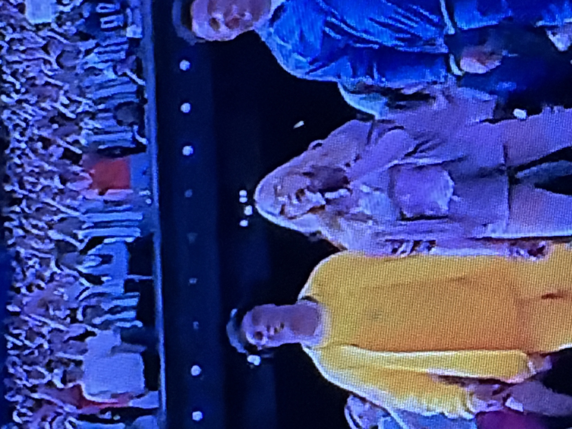 In this low-resolution, grainy image, three individuals are visible in the foreground, with a crowd of spectators in the background. The person on the left is wearing a bright yellow hooded sweatshirt. To their right, a person with blonde hair is wearing a light purple outfit that appears to be a jacket over a corset-like top and pants. The person on the far right is wearing a dark blue jacket. The background shows a large crowd of people, many of whom are wearing white t-shirts or jerseys with black and white stripes, suggesting a sporting event or concert. The lighting is dim, with some bright circular lights visible above the crowd. The overall scene appears to be an outdoor event at night.