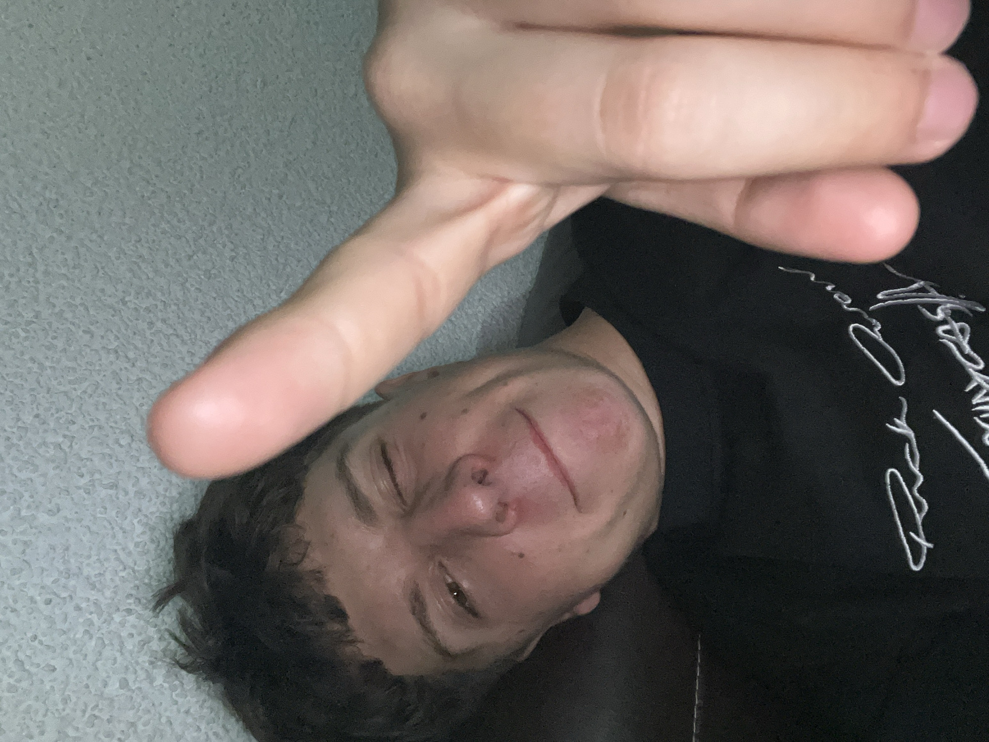 The image depicts a close-up, low-angle perspective of a young adult male, captured in Zlatograd, Bulgaria. The subject has short, dark brown, textured hair with bangs. He is winking with his right eye (viewer's left) and displays a slight smirk or smile on his lips. His left eye is open and directed towards the camera. The subject's right hand is extended into the immediate foreground, with the index finger pointing directly at the camera lens, causing it to appear large and slightly out of focus, partially obscuring the upper-right portion of the frame. He is wearing a black t-shirt with white, stylized cursive text, possibly "Rick Owens," visible on the lower-left quadrant of the garment. The background consists of a textured, light gray or muted green surface, likely a wall or ceiling, illuminated by dim, ambient lighting. Shadows are present on the left side of the subject's face (viewer's right) and under his chin. The composition suggests a self-portrait (selfie).