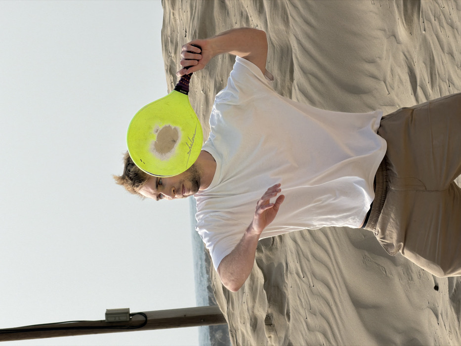 The scene depicts a single adult male in a dynamic pose on a sandy beach or dune area, consistent with the coastal regions of Tel-Aviv, Israel. The subject is a Caucasian male, approximately 20-35 years old, with short light-brown hair and light stubble. He wears a loose-fitting white short-sleeved t-shirt and light brown or khaki elastic-waisted shorts/trousers. His body is turned slightly to the right, with knees bent and feet spread in an athletic stance. His left arm is bent at the elbow, extended slightly forward. His right arm is raised, holding a neon yellow beach tennis paddle with a dark grip, positioned behind and above his head, as if mid-swing or preparing to strike. The paddle features a circular area of wear in its center, and a script-like inscription is visible on its lower face. The man's gaze is directed towards his left (viewer's left).
The environment consists of undulating sand dunes in the foreground and midground. To the far left, a tall wooden utility pole with attached dark cables and a small gray electrical box rises vertically from the sand. In the distant background, a faint horizon line suggests the presence of an ocean, likely the Mediterranean Sea. The sky is bright and appears overcast or hazy, providing diffuse illumination without harsh shadows.