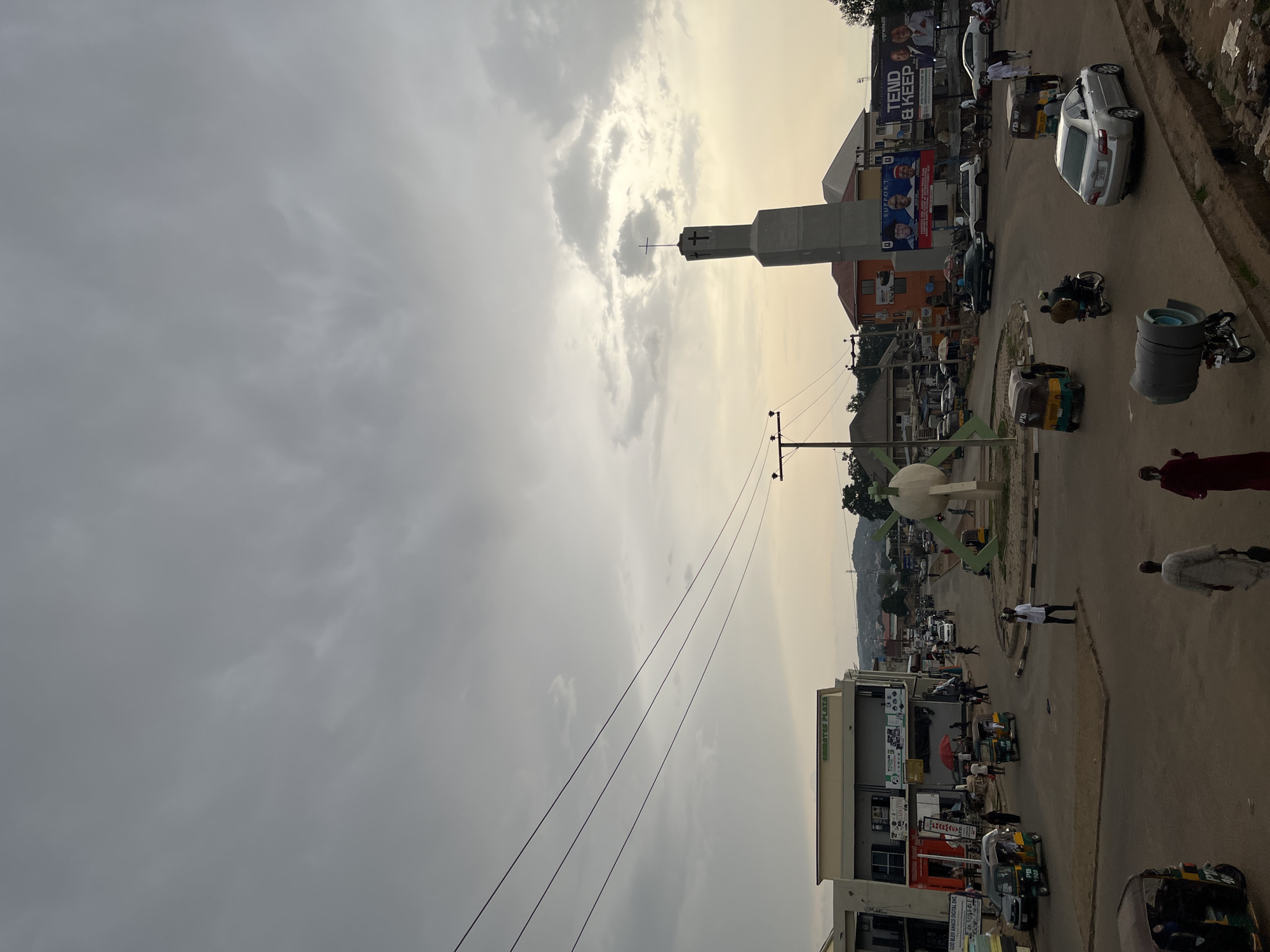 The image shows a street scene in Jos, Nigeria, with a prominent circular intersection. A concrete sculpture, possibly a monument, stands in the center of the roundabout. To the right, a tall, modern church building with a cross at its apex dominates the skyline. Various vehicles, including cars, motorcycles, and auto-rickshaws (keke NAPEPs), are visible on the road. Several individuals are walking along the street and sidewalks. Buildings line the sides of the road, featuring shops and businesses, with signage like "EMIRATES PLAZA" visible. Billboards advertising political figures and campaigns are also present. The sky is overcast with heavy clouds, suggesting the possibility of rain or an approaching storm. The overall atmosphere appears to be that of a bustling urban environment.