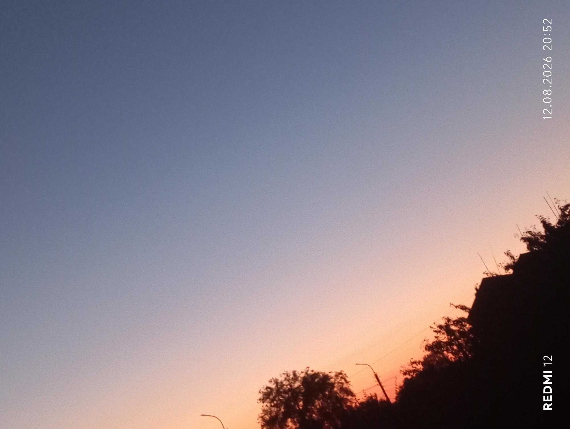 The image is a photograph of the sky at dusk, with a gradient of colors from orange near the horizon to blue higher up. Silhouetted against the sky are trees, some power lines, and a building. The bottom left corner of the image displays "REDMI 12" in white text, and the bottom right corner shows "12.08.2026 20:52" in white text. The image was taken in Smila, Ukraine.