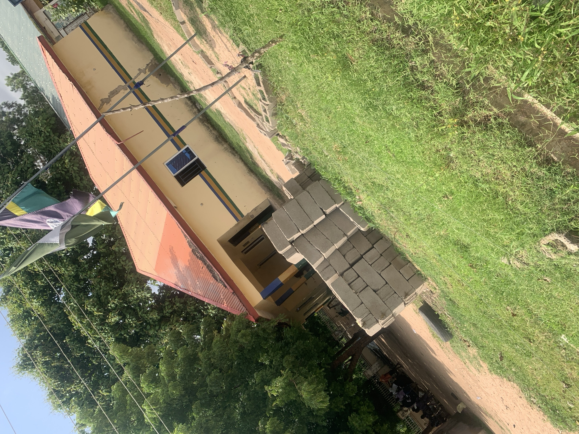 The image shows the exterior of a single-story building, likely a school or government office, located in Jalingo, Nigeria. The building has a light yellow facade with blue, green, and yellow horizontal stripes running across it. The roof is orange and appears to be corrugated metal. Three flags, one green and two purple/yellow striped, are planted in front of the building, attached to metal poles. A stack of grey concrete blocks is visible in the foreground, positioned on a patch of grass and dirt. Behind the blocks, a discarded car tire lies on the ground. The area around the building is partially paved with dirt and partially covered in grass. Several trees with dense green foliage are situated to the left of the building. In the background, to the left of the main building, a group of approximately 5-7 people are gathered, some seated on benches under a tree. The sky is visible and appears to be clear and blue. The overall scene suggests an outdoor, possibly construction or maintenance, area adjacent to a public building in a tropical or sub-tropical climate.