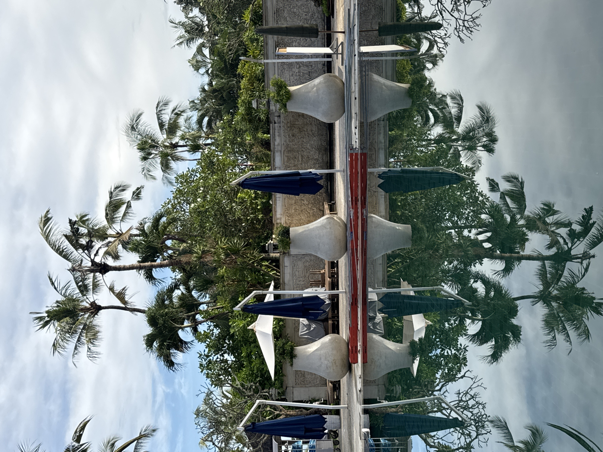 This image depicts a serene, tropical outdoor setting, likely a resort or hotel area in Kuta Selatan, Indonesia. The scene is dominated by a long, horizontal body of water, possibly a swimming pool or a decorative pond, that creates a strong reflection of the surroundings.
In the upper half of the image, representing the real environment, a partly cloudy sky with patches of blue is visible, particularly on the left. Lush green tropical vegetation, including numerous tall palm trees with swaying fronds, borders the scene. Along a structured pathway or low wall, several large, light-colored, decorative urns or planters are spaced out, some featuring vibrant green foliage. Blue and white patio umbrellas, mostly closed, are positioned beside these structures. Red and silver architectural elements, possibly parts of railings or furniture, are also present.
The lower half of the image showcases a near-perfect, yet slightly muted and darker, reflection of the upper scene in the water. The sky, palm trees, urns, umbrellas, and structural elements are all mirrored with remarkable clarity, creating a striking symmetrical composition.
No people are visible, indicating a quiet and unoccupied leisure space. The time of day appears to be daytime, under a bright, partly cloudy sky. The tropical flora and architectural style are consistent with the provided location context of Kuta Selatan, Indonesia. There is no visible text in the image.