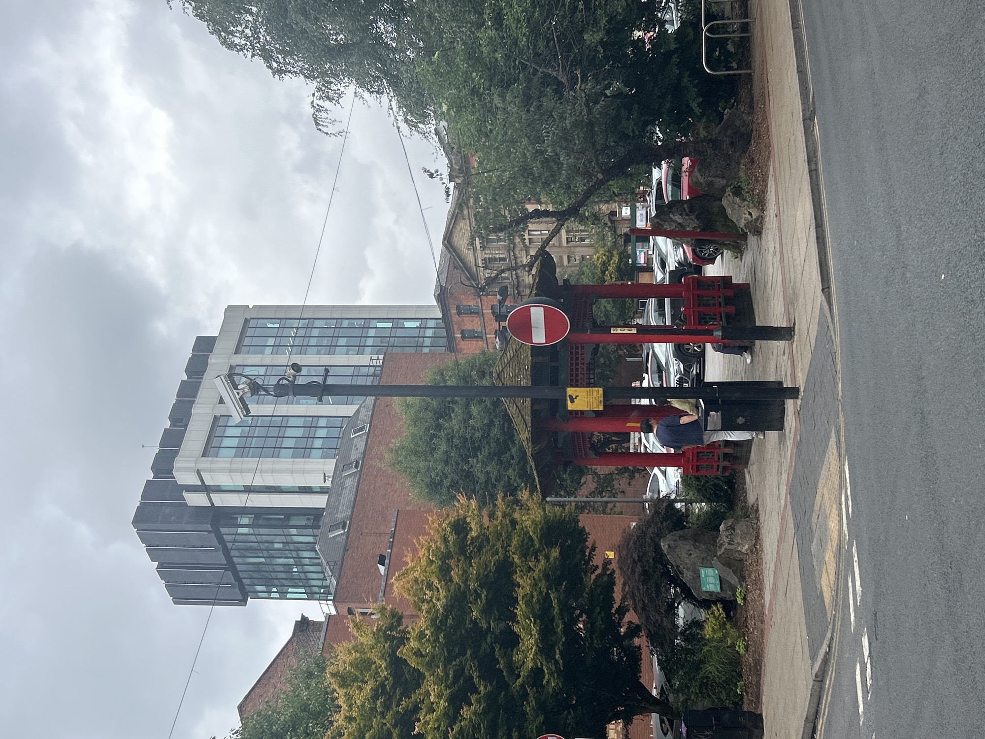 This image depicts the exterior of the Manchester Chinese Centre on Nicholas Street in Manchester, United Kingdom. The building features a distinctive red, pagoda-style entrance with ornate detailing. In front of the centre, a large tree with a multi-colored canopy is visible, along with several large rocks and manicured landscaping. A lamppost with a CCTV camera and a "no entry" sign is positioned near the entrance. A person in a dark blue shirt and light-colored pants stands near the centre. The background shows a mix of modern and older architecture, including a tall, glass-fronted building and a red brick building. The sky is overcast. The scene is set on a street with a pedestrian walkway and visible road markings, suggesting an urban environment.