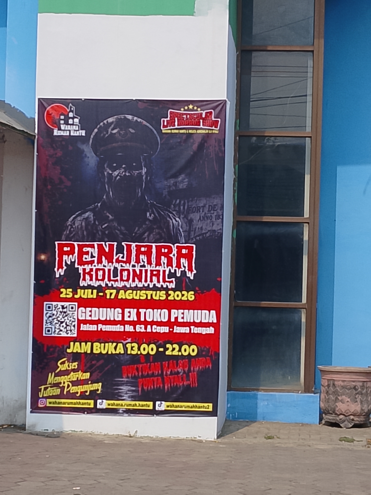 A vertical banner displays a spooky Halloween-themed attraction. At the top left, a logo depicts a red moon behind a haunted house and the text "WAHANA RUMAH HANTU." The central image is a large, terrifying figure in a military-style hat and uniform, with glowing white eyes and a skeletal face. The banner's title, "PENJARA KOLONIAL," is written in large, distressed red letters. Below this, event details are listed: "25 JULI - 17 AGUSTUS 2026," followed by "GEDUNG EX TOKO PEMUDA" and the address "Jalan Pemuda No. 63. A Cepu - Jawa Tengah." A QR code is prominently displayed. The operating hours are "JAM BUKA 13.00 - 22.00." A slogan reads, "Sukses Menggetarkan Jutaan Pengunjung," with accompanying social media handles: "wahanarumahhantu" on Instagram and TikTok. The bottom of the banner features the text "BUKTIKAN KALAU ANDA PUNYA NYALI!" and a TikTok logo. The banner is attached to a white wall, with a blue wall and a window visible to the right. A decorative ceramic pot sits on the ground to the far right. The ground appears to be paved with interlocking bricks. The location is implied to be in the city of Ngelo, Indonesia, given the context of the text on the banner.