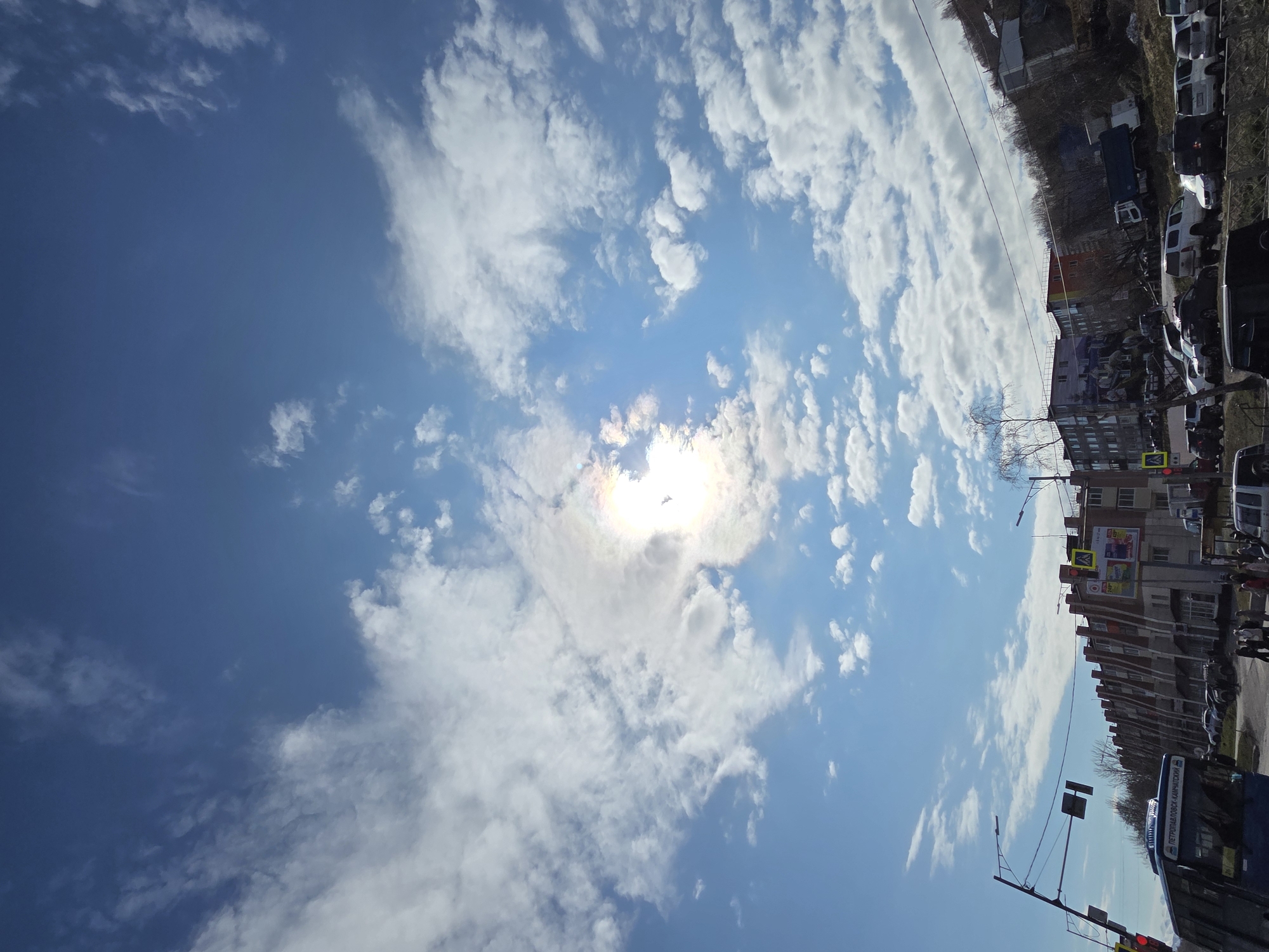 The image depicts a partly cloudy sky above a street scene in an urban environment. The sun is visible through a thin layer of clouds in the center of the frame. The street is lined with buildings of varying heights and architectural styles, including residential and commercial properties. Parked cars are visible along the street. A pedestrian crossing sign and traffic lights are visible at an intersection. A blue bus with Cyrillic script on its side is present. A few bare trees are scattered amongst the buildings.