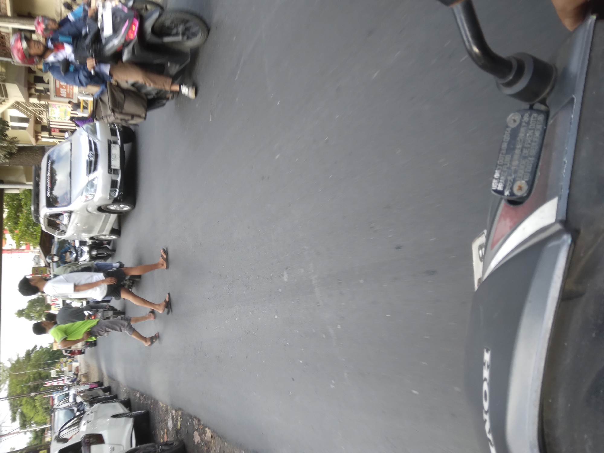 The image captures a street scene in Desa Pamijen, Kecamatan Sokaraja, Indonesia, focusing on a moment of pedestrian and vehicular activity. In the foreground, the edge of a dark-colored scooter is visible, with the "HONDA" logo imprinted on its body. A warning label regarding brake fluid is also present on the scooter.
On the road, two men are crossing. One is wearing a green t-shirt and grey shorts, while the other is in a white t-shirt and dark shorts. Both are wearing flip-flops. Behind them, a silver van is stopped, likely yielding to the pedestrians. Further down the road, another silver van, this one with tinted windows, is in motion. To the right, a motorcyclist wearing a red helmet and a blue jacket is riding a scooter. The road surface is asphalt, and the surroundings show some trees and buildings, suggesting an urban or semi-urban setting. The lighting indicates it is daytime.