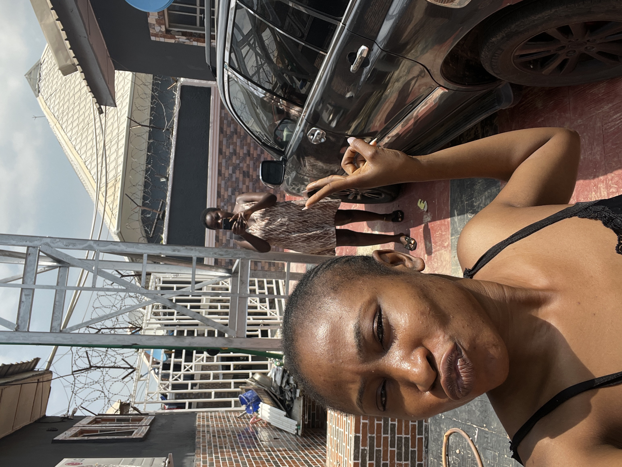 In an outdoor courtyard in Abuja, Nigeria, a woman in the foreground is taking a selfie, pouting her lips. Behind her, another woman stands near a dark SUV, holding a mobile phone and appearing to take a photo. The environment includes a brick wall, a metal structure, a white gate with barbed wire on top, and a tiled floor. The lighting suggests it is daytime.