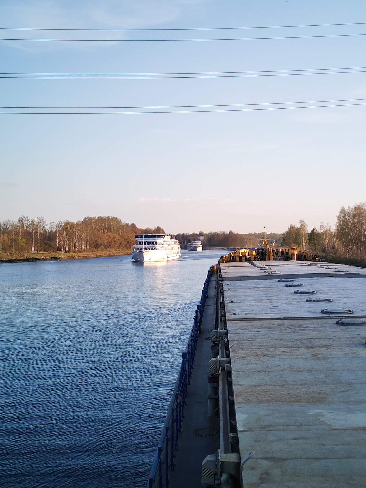 The image shows a waterway in Базарово, Russia. In the middle ground, a white passenger cruise ship is moving on the water. Further in the distance, another vessel is visible. The foreground is dominated by the flat, gray deck of a barge. On the sides of the waterway are trees, and power lines stretch across the sky. The sky is a pale blue with a slight gradient.