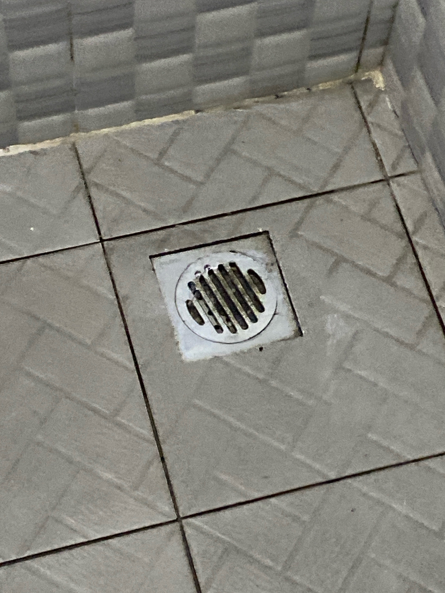 The image shows a close-up, high-angle shot of a square shower drain with a circular grate on a tiled floor. The tiles are laid in a herringbone pattern. The drain and surrounding grout appear to have some discoloration and possibly mildew. The background shows a tiled wall with a horizontally aligned pattern. The location is identified as Klurak, Indonesia.