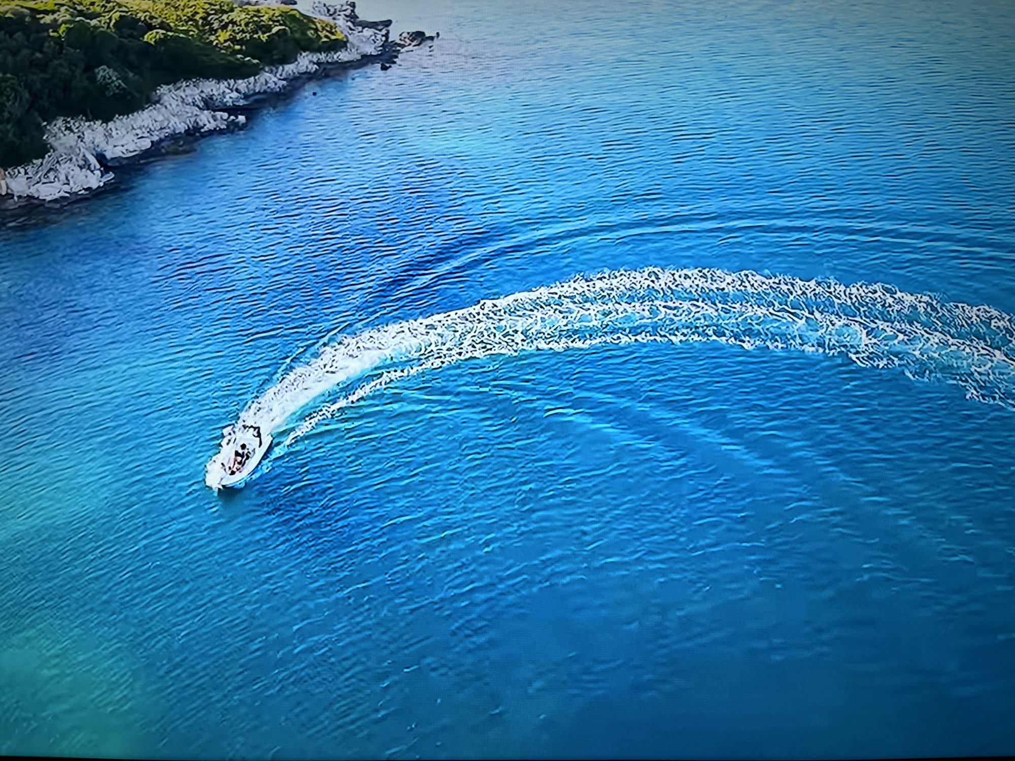 The aerial view captures a white speedboat with several passengers navigating through clear blue water. The boat is leaving a white wake as it turns, with the wake fanning out behind it. To the left of the boat, a rocky coastline is visible, covered in green vegetation. The water exhibits ripples and varying shades of blue, suggesting depth and clarity. The scene depicts a leisurely activity in a natural, coastal environment. The provided image does not contain any visual cues or landmarks that would allow for the identification of the city of Kharkiv, Ukraine.