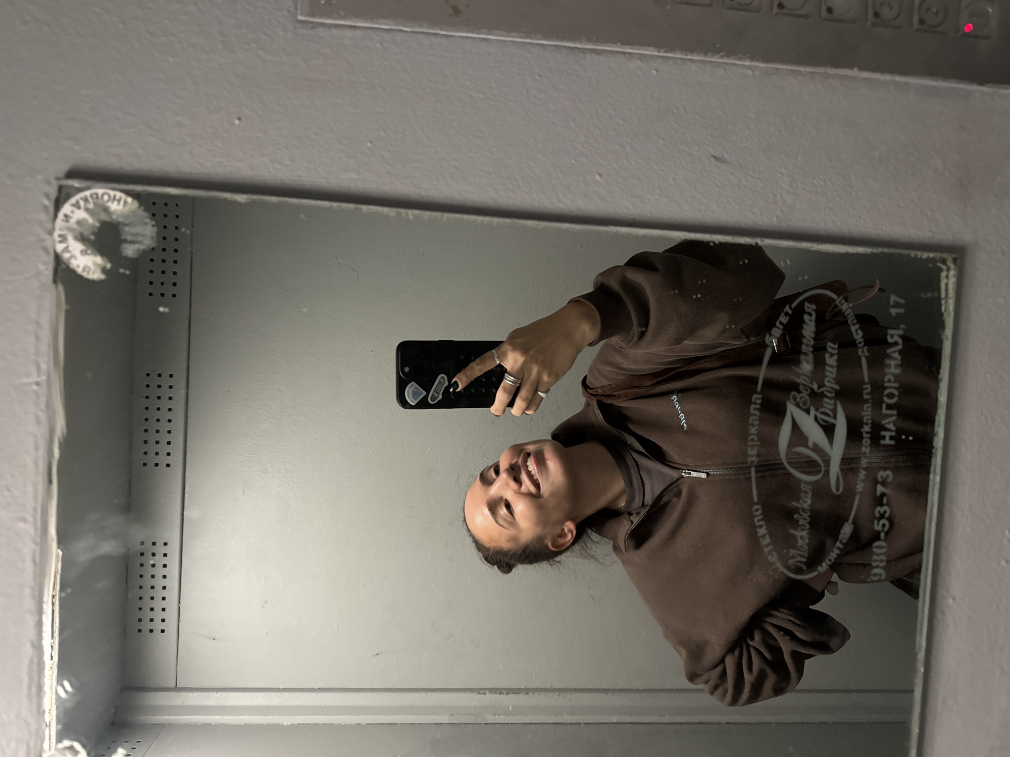 The image depicts a woman taking a selfie in a mirror. The mirror has text and a logo printed on it, indicating it's manufactured by a glass and mirror company called "Moskovskaya Fabrika Zerkal" (Moscow Mirror Factory). The company's website is "www.zerkala.ru", and their phone number is 980-53-73. Their address in Moscow is "Nagornaya, 17" (Улица Нагорная, дом 17). The woman is wearing a brown zip-up sweatshirt. The environment appears to be an elevator, due to the mirrored surface, and the perforated metal panel visible in the upper left corner.