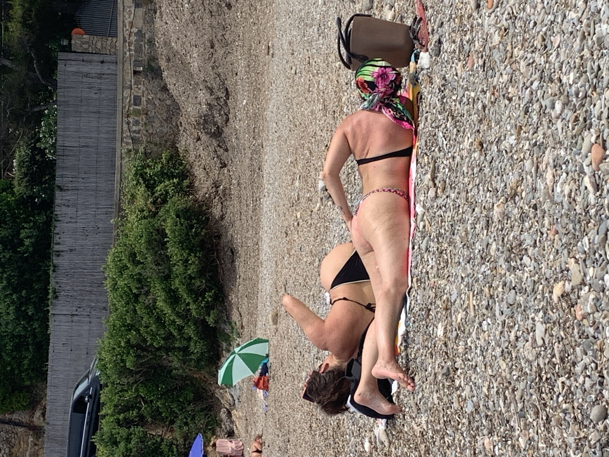 In Pozzarello, Italy, two individuals are sunbathing on a pebbled beach. The person in the foreground, lying on their stomach, has their legs extended and is wearing a black bikini bottom and a colorful headscarf. To their left, another person is lying on their stomach, facing away from the camera, wearing a black bikini. A dark-colored bag is placed near the person with the headscarf. Behind them, a green and white umbrella is partially visible, along with other beach accessories. The background features dense green foliage, a wooden fence, and a glimpse of a vehicle.