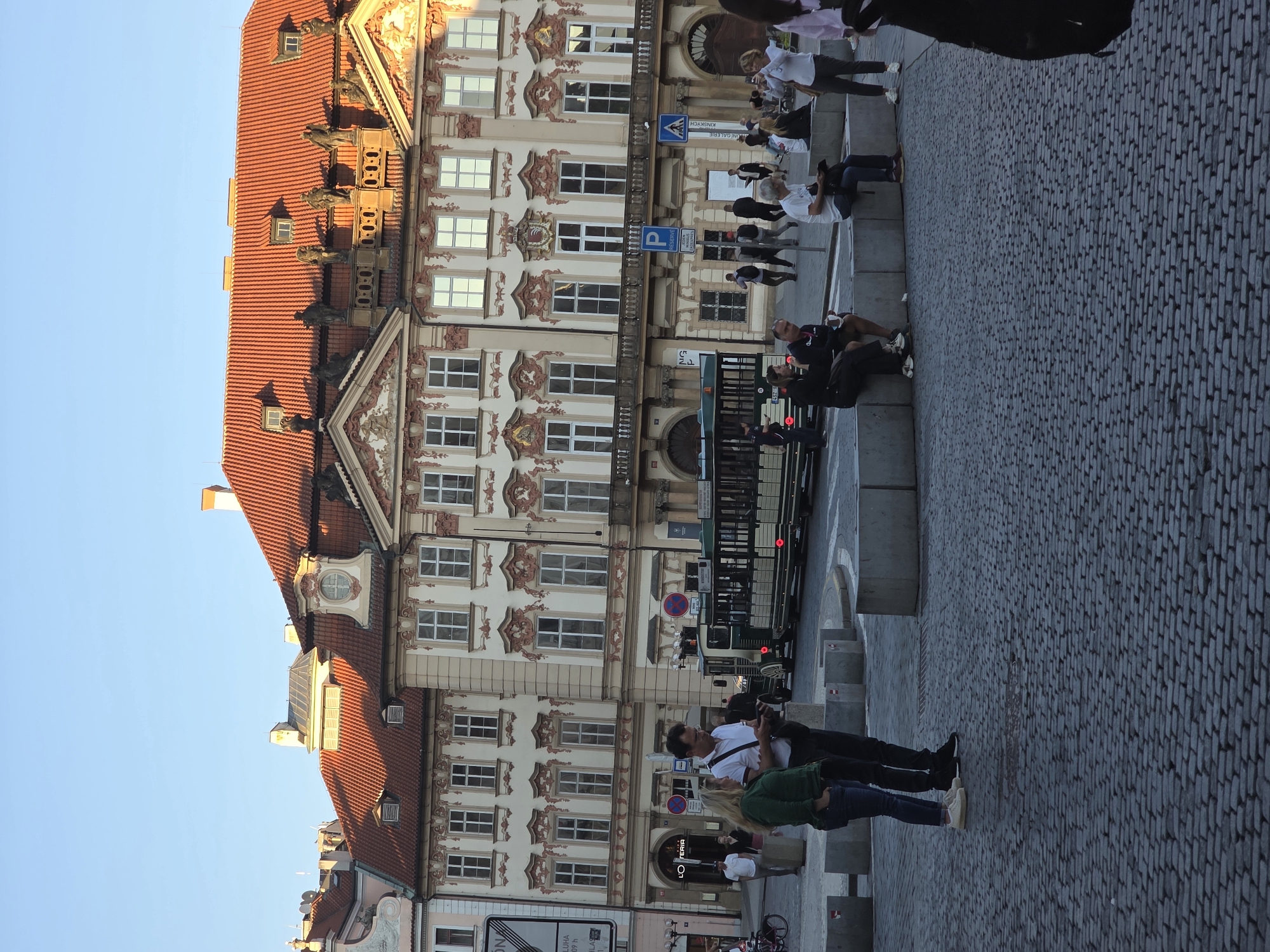 The image captures a vibrant street scene in a historic European city, likely Prague, under a clear blue sky, suggesting late afternoon or early evening. The main subject is a grand, multi-story building dominating the left side, featuring a distinctive red tiled roof with multiple dormer windows and ornate gargoyle-like figures protruding from the eaves. Its light-colored facade is richly adorned with intricate Baroque or Rococo architectural details, including decorative stucco work, classical columns, and sculptures around the windows.
In the foreground, a wide cobbled street or square, paved with grey, rectangular stones, stretches across the scene. Along the right side of the street are low, light-colored stone barriers or benches, where several people are seated, resting or observing the surroundings. Other pedestrians are seen walking and standing on the pavement. A person in a green jacket and blue jeans sits prominently on one of these barriers, conversing with another individual in a white shirt and dark pants standing beside them. Further back, more people are visible near what appears to be a tram stop, where a dark-colored tram or bus with red lights is partially visible.
The scene depicts typical urban activity, with people casually going about their day. The warm sunlight bathes the upper parts of the building, casting shadows across the street.
Several texts are visible:
* A blue sign clearly reads "KINSKÝCH GALERIE," indicating the Kinský Gallery, a notable landmark in Prague.
* Another sign shows "P RESERVOVANÉ," likely a parking sign for reserved parking.
* A shopfront further down the street displays "L'OSTERIA," a restaurant.
* Parts of other signs include "LOUBNA" and "NOVA."