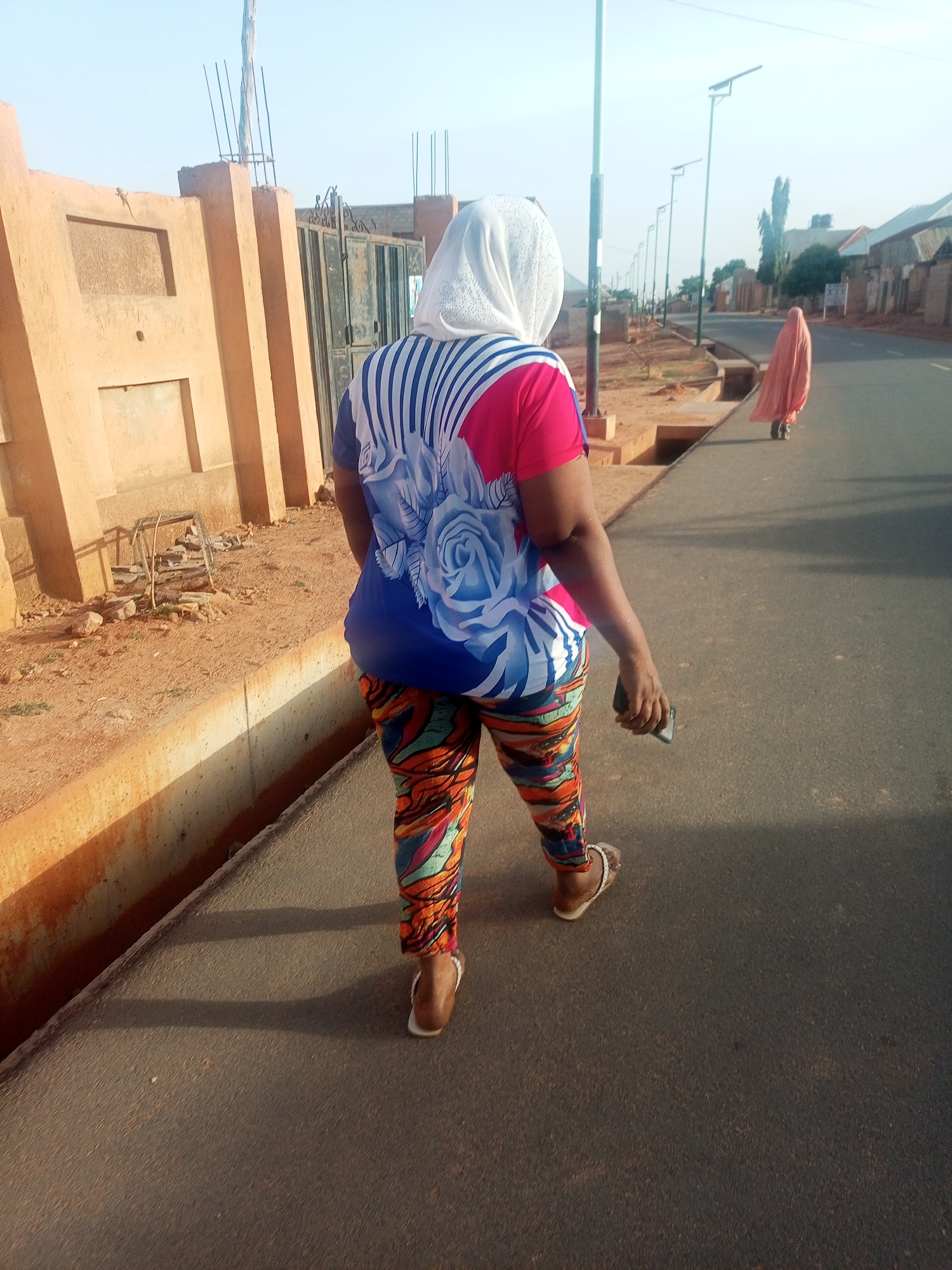 A woman in a headscarf is walking down a paved road. She is wearing a short-sleeved shirt with a rose print and patterned pants and sandals. She holds a mobile phone in her right hand. Another person in a peach-colored covering walks further down the road. The road is lined with buildings, walls, and light posts. A concrete curb runs alongside the road. The sky is light blue.
