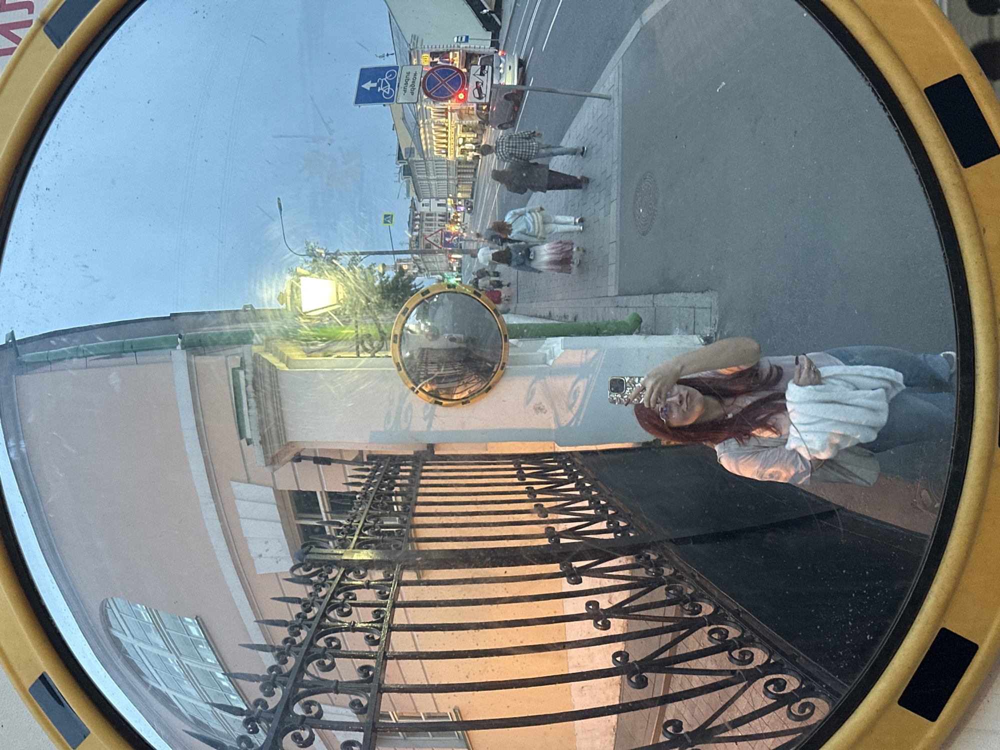 Captured from a round mirror with a yellow frame, the scene presents a street in Unknown, Unknown. The right side of the reflection showcases a sidewalk with pedestrians walking in both directions, framed by street signs and buildings in the background. On the left, a wrought iron fence lines a building with several windows. In the foreground, the mirror reflects a woman taking a photo with her phone. The overall light suggests it is daytime, or perhaps dusk, illuminating the details within the mirror.