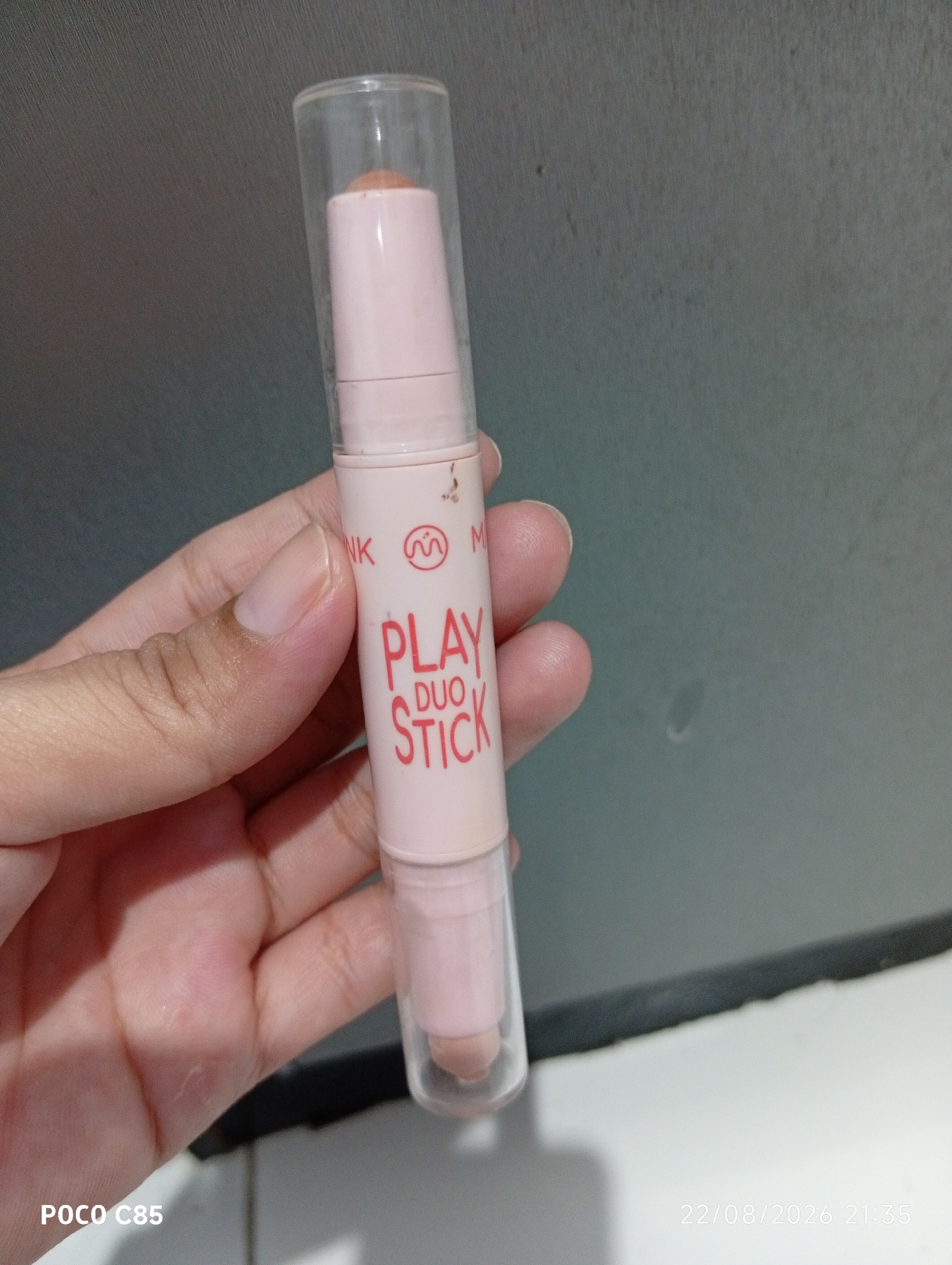 A person's left hand holds a pink makeup stick with a clear plastic cap. The makeup stick has red text on it that reads "PLAY DUO STICK". The background is a dark gray wall. A timestamp in the lower right corner reads "22/08/2026 21:35". A "POCO C85" watermark is in the lower left corner. The scene appears to be indoors.