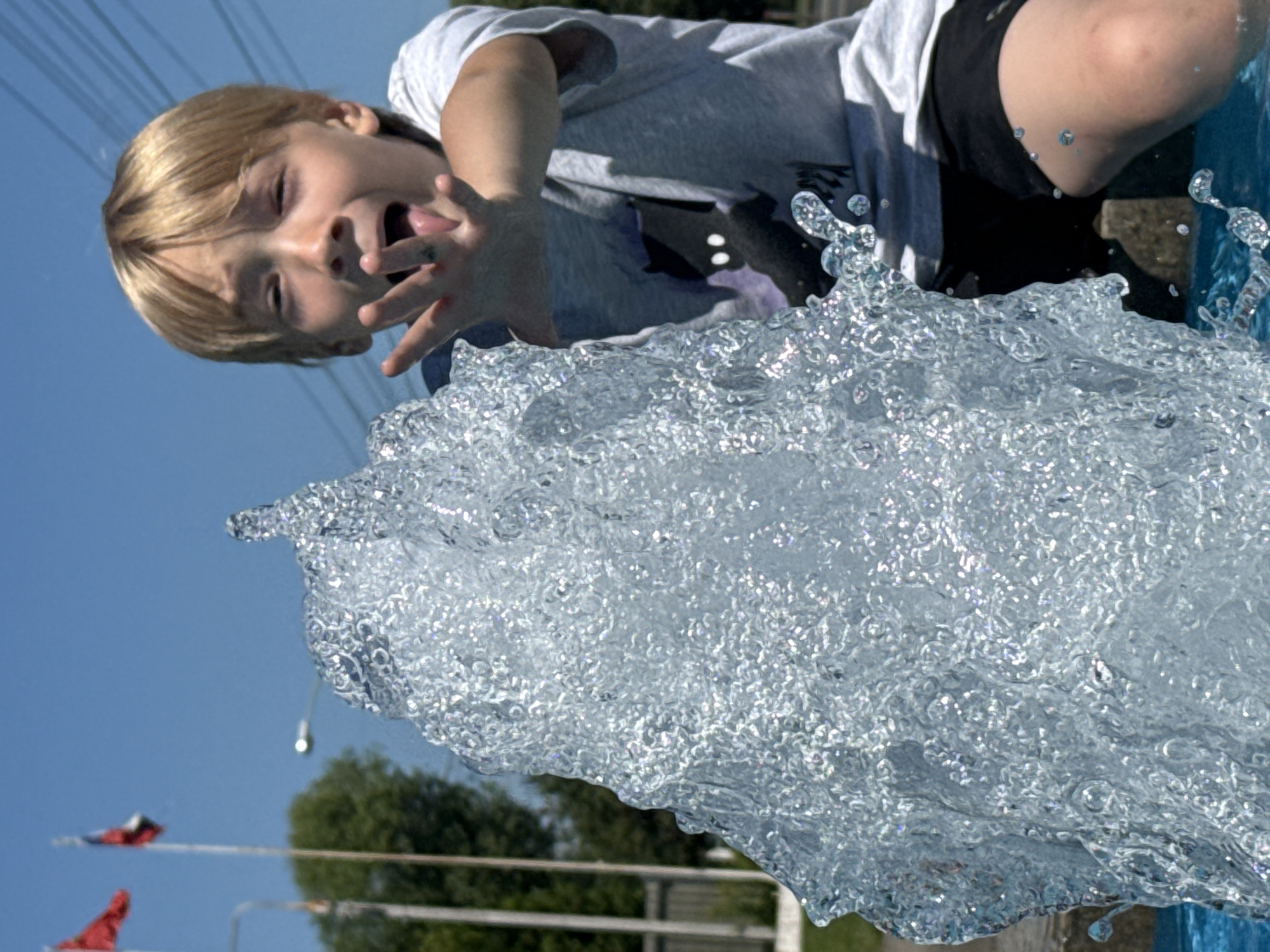A young boy with blonde hair is playfully sticking his tongue out while reaching towards a splashing water fountain in Dmitrov, Russia. He is wearing a grey t-shirt with a dark graphic print and black shorts. The fountain is actively spraying water, creating a translucent, bubbly effect in the foreground. In the background, two flags, one red and one red and white, are visible on poles against a clear blue sky. Trees and a street light are also discernible in the distance, suggesting an outdoor, public location within the city. The overall impression is one of energetic, summer fun.