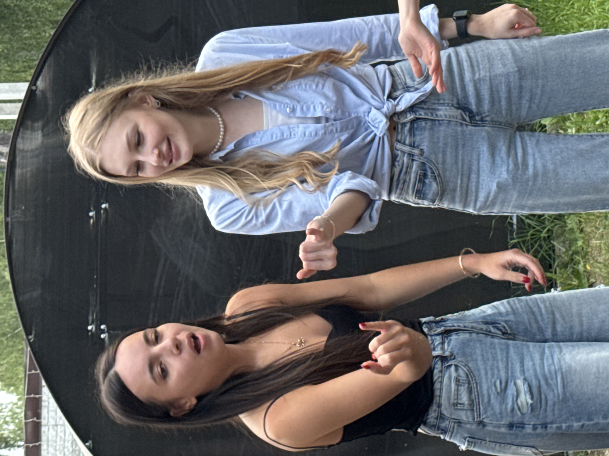 Two young women are captured in a candid outdoor shot, appearing to be in motion or mid-conversation. The image is oriented vertically, with the women positioned from top to bottom in the frame.
The woman on top has long, blonde hair and is wearing a light blue, unbuttoned shirt tied at the waist, revealing a white undergarment. She also wears a pearl necklace and light-wash denim jeans. Her expression is relaxed and joyful, with a slight smile. Her hands are gesturing outwards.
The woman below has dark, long hair and is wearing a black tank top with thin straps, adorned with a delicate gold necklace. She is also wearing light-wash denim jeans, with some distressing. Her mouth is open, suggesting she is speaking or reacting to something. Her hands are also in a gesturing pose.
Both women are dressed in casual, summery attire. The background is divided. To the left, a dark, curved surface dominates, possibly a satellite dish or a similar structure, with some metallic fixtures visible. To the right and behind the women, there is lush green grass and some foliage, suggesting an outdoor setting like a park or a backyard. The lighting suggests it is daytime, possibly late afternoon or early evening, with soft shadows. The overall mood is lively and informal, hinting at a moment of fun or energetic interaction between the two friends. There is no visible text in the image. The context provided places this scene in Perm, Russia.
