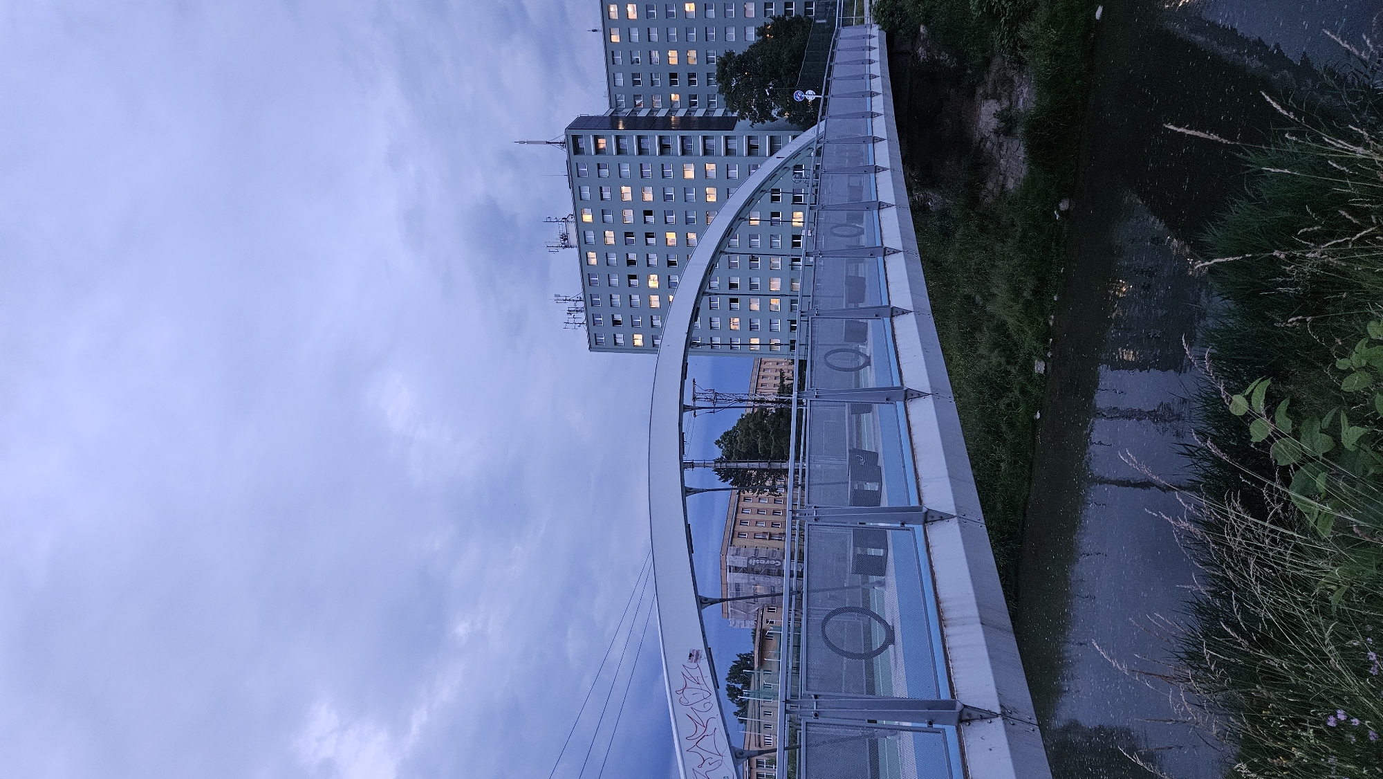 The image captures a pedestrian bridge arching over a narrow, dark river in the city of Prešov, Slovakia, likely in the Sekčov district given the presence of socialist-era residential buildings. The sky is overcast with gray clouds, suggesting either dusk or an impending storm. The bridge itself is modern, with a white arched structure and metal railings. The bridge deck is painted blue, and there are what appear to be benches and decorative circular elements on its surface. Several large, block-like apartment buildings, characteristic of the socialist era, dominate the background. These buildings are lit from within, indicating that people are inside. The riverbank on the right side of the image is overgrown with grass and reeds, while the left bank is also vegetated, with some scattered rocks visible. Power lines are visible in the upper left portion of the image, and there is graffiti on the side of the bridge's arch. The overall atmosphere is somewhat somber and urban.