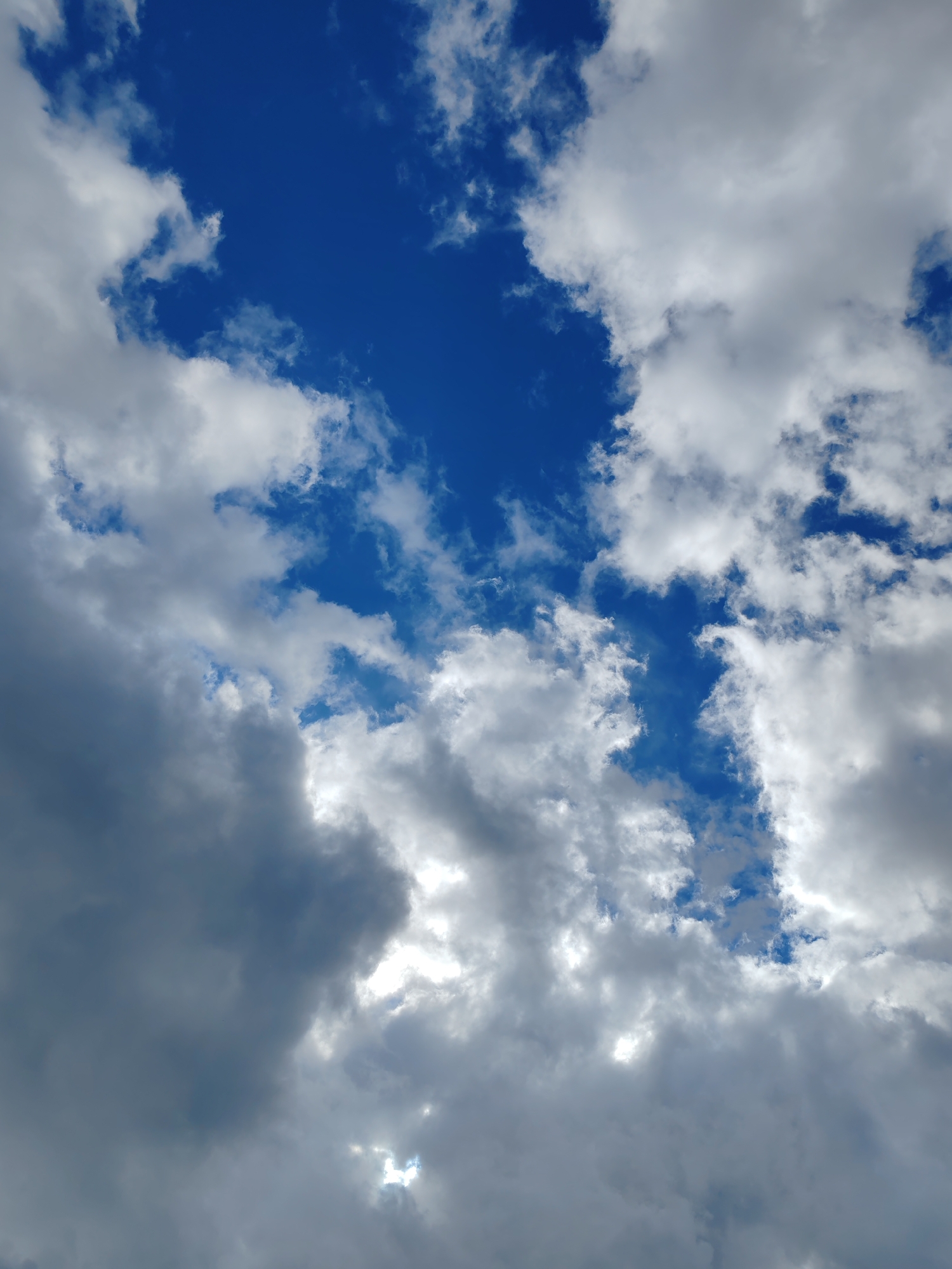 The image displays a dynamic sky filled with a mix of dark, heavy clouds and bright, luminous patches of blue sky peeking through. Sunlight filters through the clouds, creating areas of bright illumination and casting shadows. The perspective is looking upwards, capturing the vastness of the sky. The scene appears to be an outdoor natural environment, typical of the weather conditions that might be experienced in Чухлэм, Russia, a city known for its variable climate. There are no people, specific objects, or actions depicted in the image; it is solely a natural atmospheric scene.