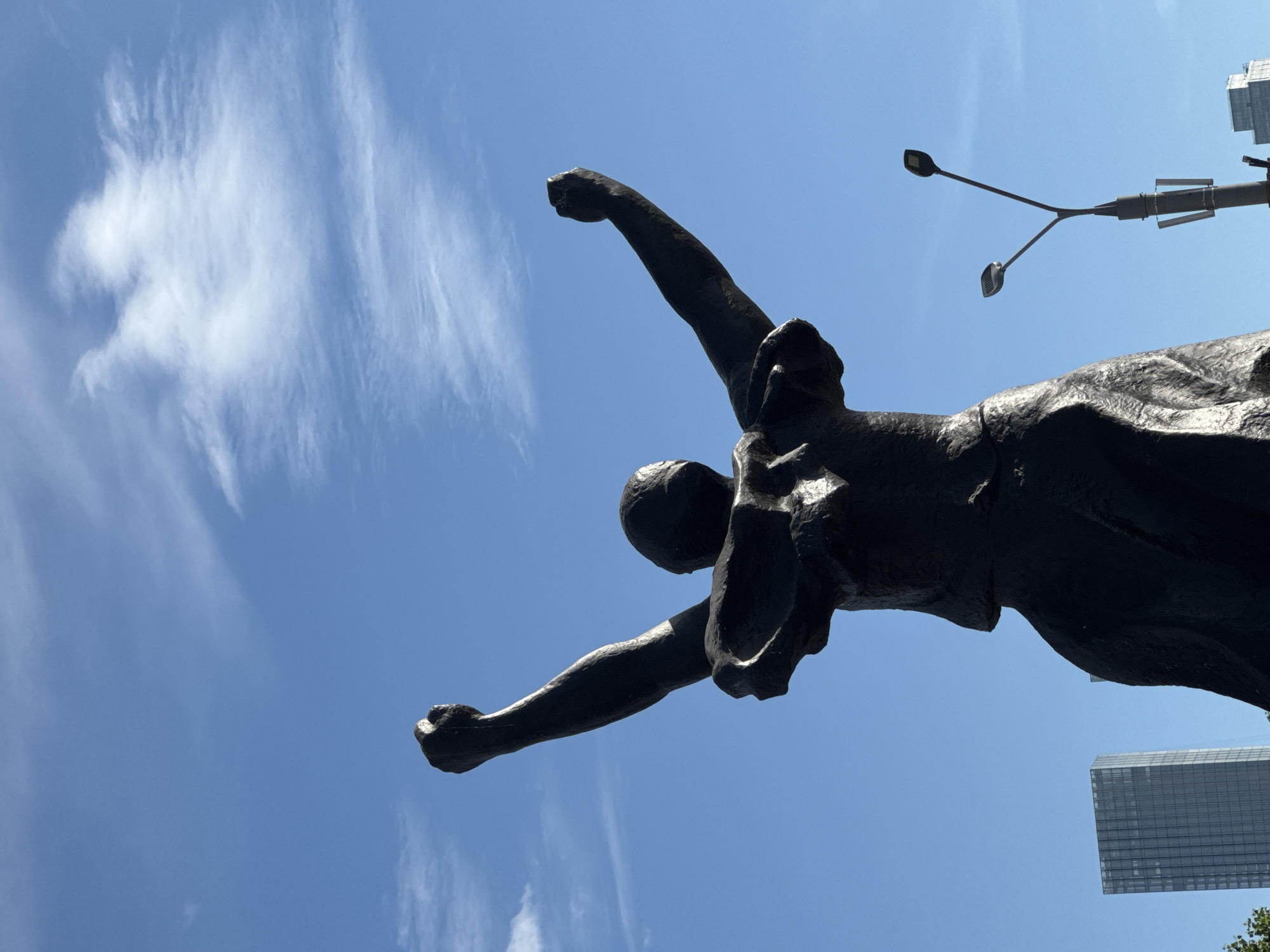 A dark-colored, full-body statue of a person with arms raised in triumph stands against a clear blue sky with scattered white clouds. The statue's head is obscured, and its chest is depicted with folds, suggesting clothing. The figure is wearing a dress that drapes around its lower body. In the background, a modern, tall, glass-paneled skyscraper is visible on the left, and a streetlamp with two lights rises on the right. The statue is located in Moscow, Russia, specifically on Bolotnaya Square, near the monument to Peter the Great.