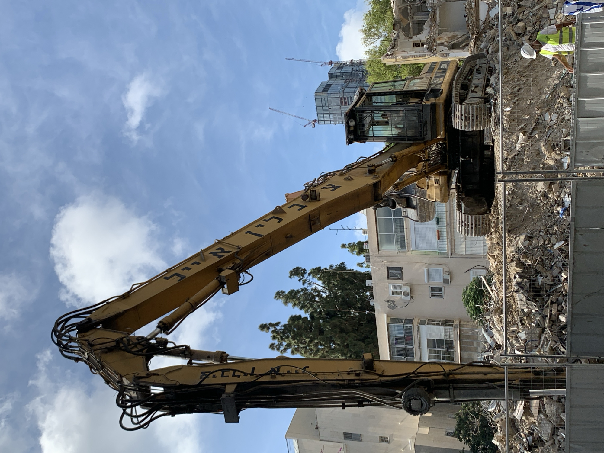 A large, yellow excavator, with text in Hebrew script on its arm, stands amidst a construction or demolition site in an urban environment. The excavator is positioned on a pile of rubble, likely from a building that has been torn down. In the background, there are residential buildings, some of which have air conditioning units on their exterior walls. Further back, there is a construction site with cranes. A worker wearing a white hard hat and a yellow reflective vest can be seen near a security fence. The sky is blue with scattered clouds. The general location appears to be Tel Aviv, Israel, based on the construction style and the Hebrew text on the machinery.