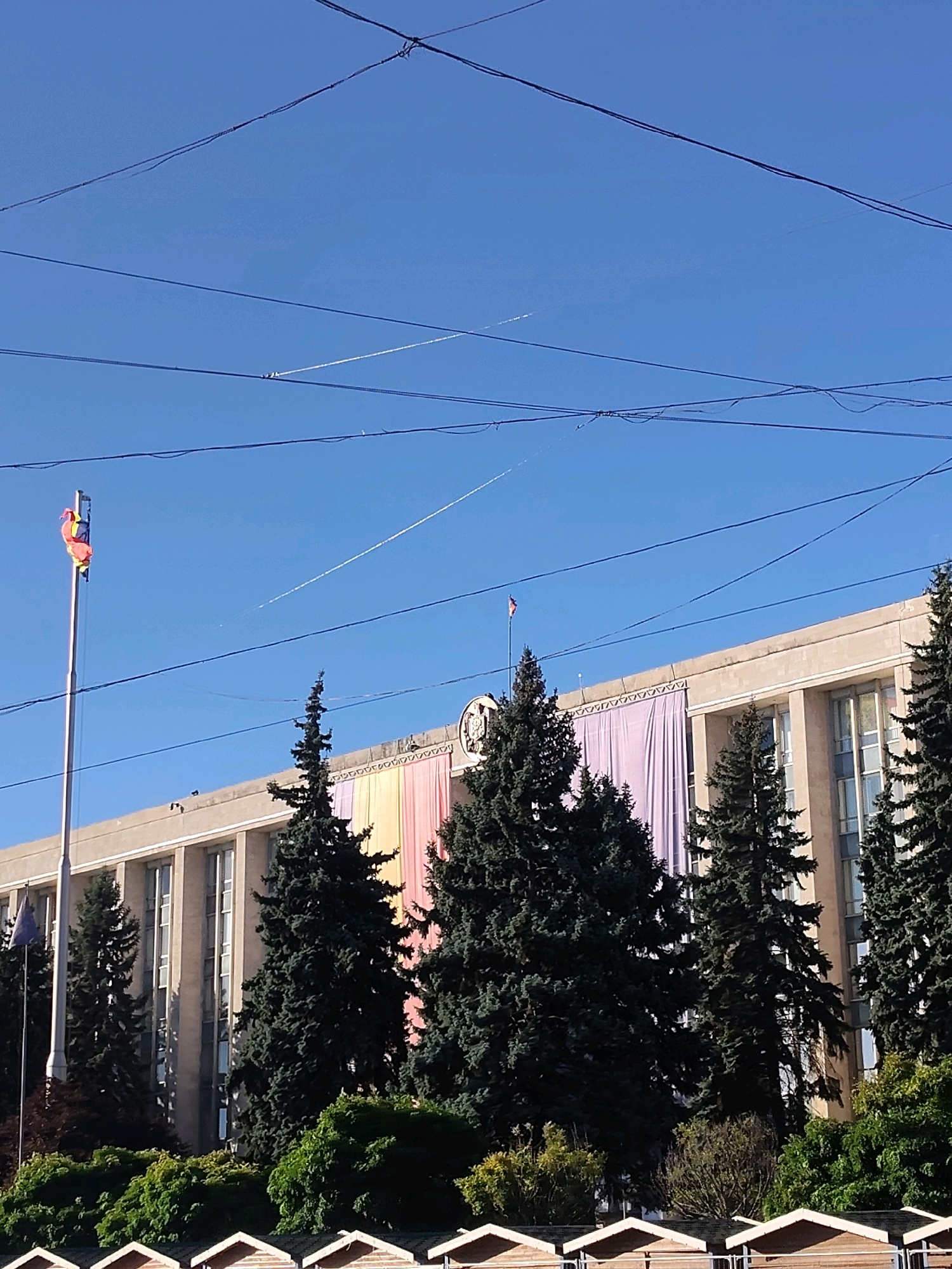 The image shows a wide shot of a large, modern-looking building with tall windows. The building is adorned with large, colorful fabric panels hanging from its facade, displaying a gradient of pastel colors, including shades of purple, pink, yellow, and orange. In front of the building, several tall, dark green pine trees stand prominently. To the left of the building, a flagpole with multiple flags is visible, one of which appears to be partially furled. The sky above is a clear, bright blue, with several thin, white contrails from airplanes crisscrossing it. Overhead, a network of black wires, possibly power or utility lines, stretches across the frame. In the foreground, the roofs of several small, wooden structures with peaked tops are visible, suggesting a street-level perspective. The overall impression is one of a public or institutional building, possibly decorated for an event, on a sunny day.