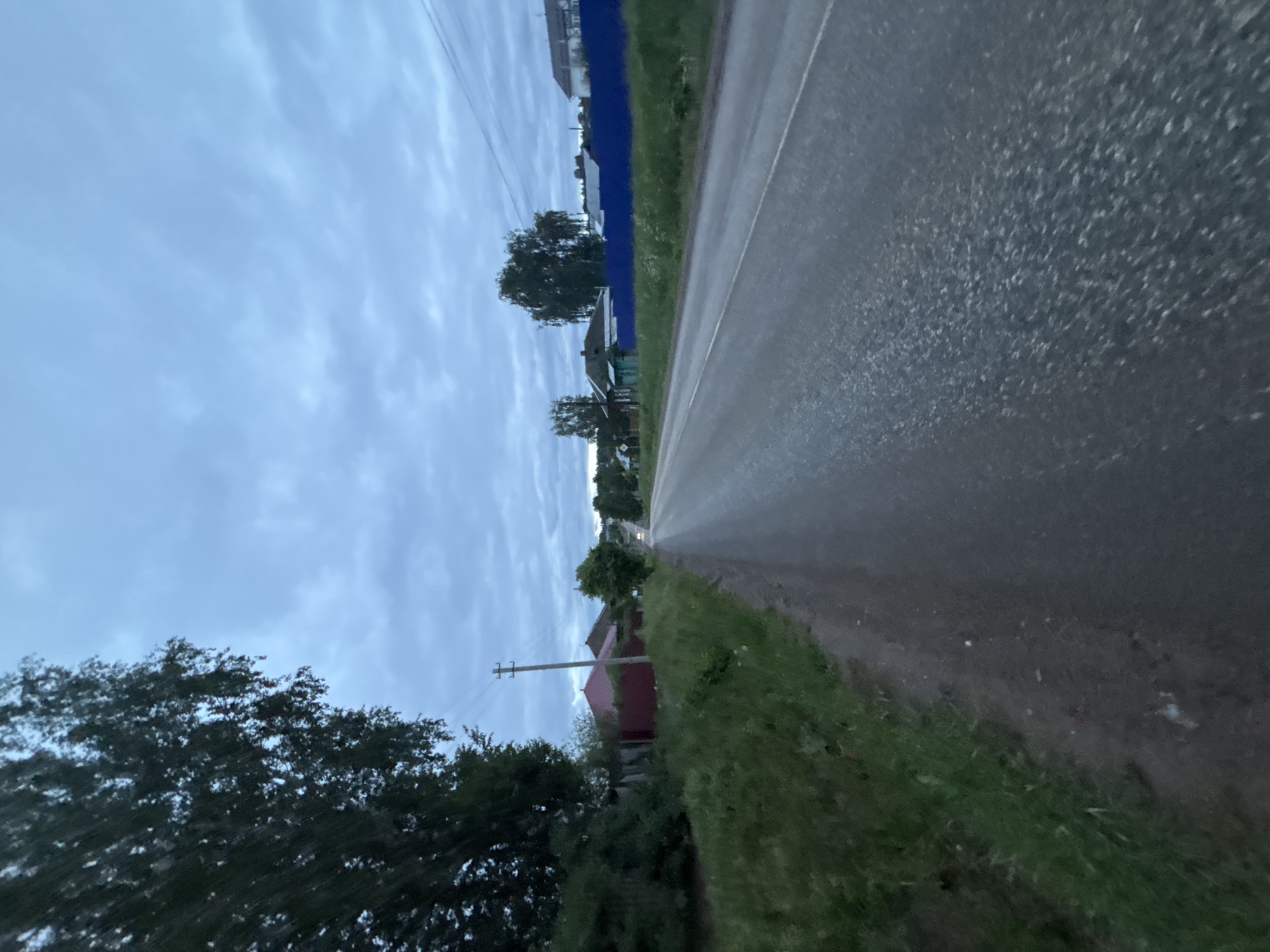 The image shows a road in Фоки, Russia. The road surface is asphalt, appearing slightly wet. On the right side of the road is grassy verge. The left side includes trees and foliage, along with glimpses of houses. A blue fence is visible further down the road on the left. Overhead power lines are visible against the cloudy sky.