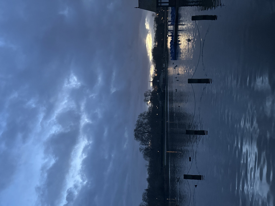 The image depicts a twilight or early evening scene at a body of water, possibly a lake or a wide river, viewed from the shoreline. The perspective is horizontally oriented, with the sky filling approximately the left third of the frame, and the water occupying the right two-thirds.
The sky is largely overcast with heavy, dark grey and blue-grey clouds. However, a significant break in the clouds along the distant horizon reveals a bright strip of light, varying from soft yellow to pale white, suggesting either a sunset/sunrise or a strong light source beyond the immediate cloud cover.
The water surface is relatively calm but shows subtle ripples, particularly on the right side, and reflects the dim light from the sky and the brighter light from the horizon. Several dark, vertical wooden posts, each with distinct white bands near the top, are spaced out in the water, extending from the foreground towards the mid-ground. These posts are connected by visible chains, forming a boundary or a decorative feature in the water.
Along the distant shoreline, silhouetted against the brighter horizon, are numerous bare trees, indicating late autumn or winter. Several distant lights, possibly streetlights or lights from buildings, dot the shoreline. A distinct building with a peaked roof is visible on the far left of the distant bank, likely a boathouse or similar structure. Further along, a tall, slender spire or church tower can be seen reaching towards the sky. Moored near the building on the left are several small boats, possibly paddle boats, appearing blue in the dim light. A number of ducks or other water birds are scattered across the water, some near the posts and chains, and others further out.
The overall atmosphere is serene and tranquil, with the low light and cloud cover creating a muted, somewhat melancholic color palette dominated by blues, greys, and dark tones. No text is visible in the image.