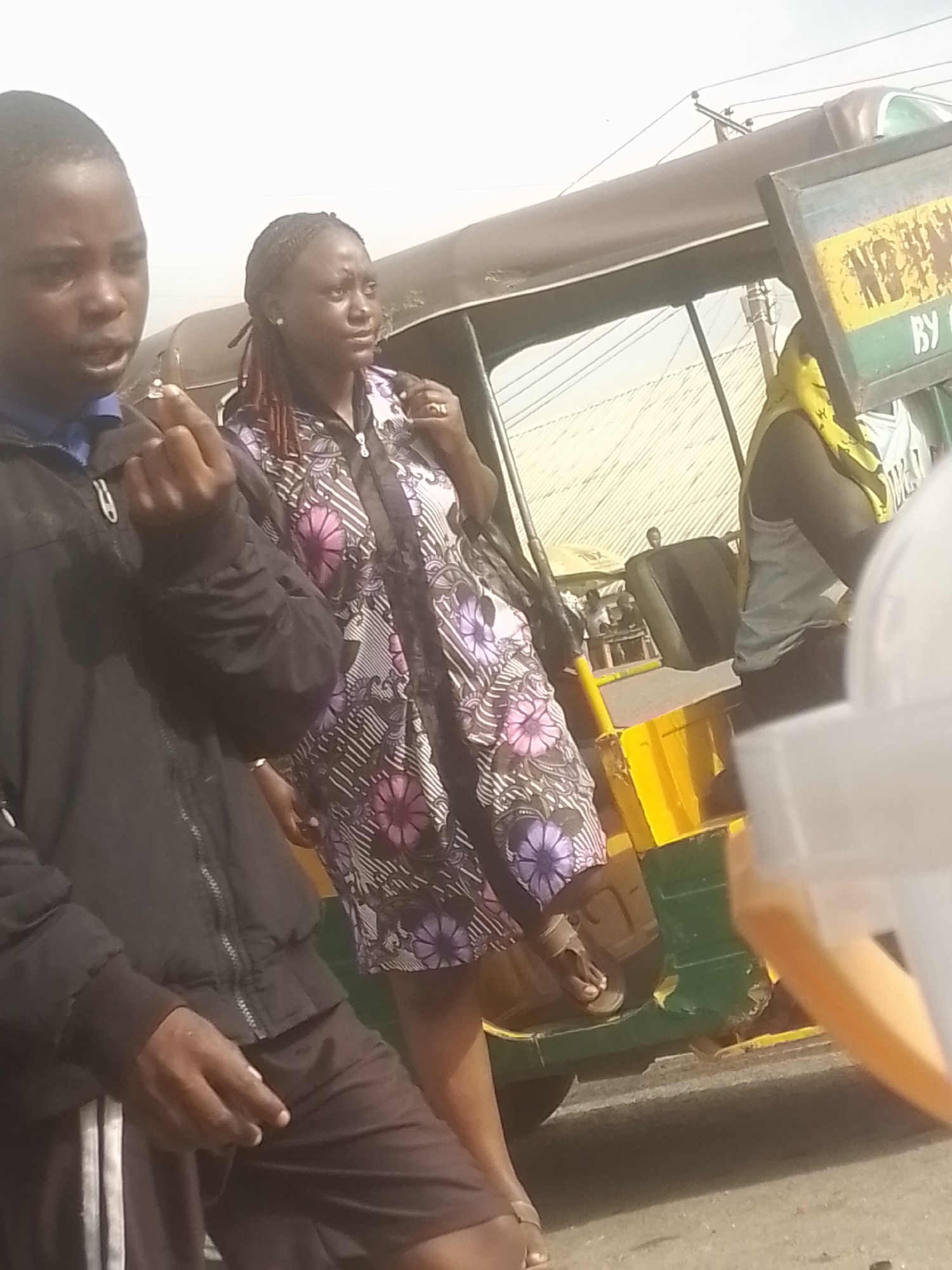 A group of people are gathered near an auto-rickshaw. In the foreground, a young person in a black jacket is looking towards the viewer and holding up an object in their right hand, possibly a cigarette. To their right, a woman in a patterned dress with purple flowers is standing and looking away. Behind them, a person wearing a yellow headscarf and a light-colored tank top is visible. The auto-rickshaw is yellow and green, with a brown roof. A sign with yellow and green panels is attached to the side of the auto-rickshaw, with some illegible white text on it. The scene appears to be outdoors, with a bright, possibly overcast sky. The ground is paved. The overall impression is a candid street scene, possibly during the daytime.