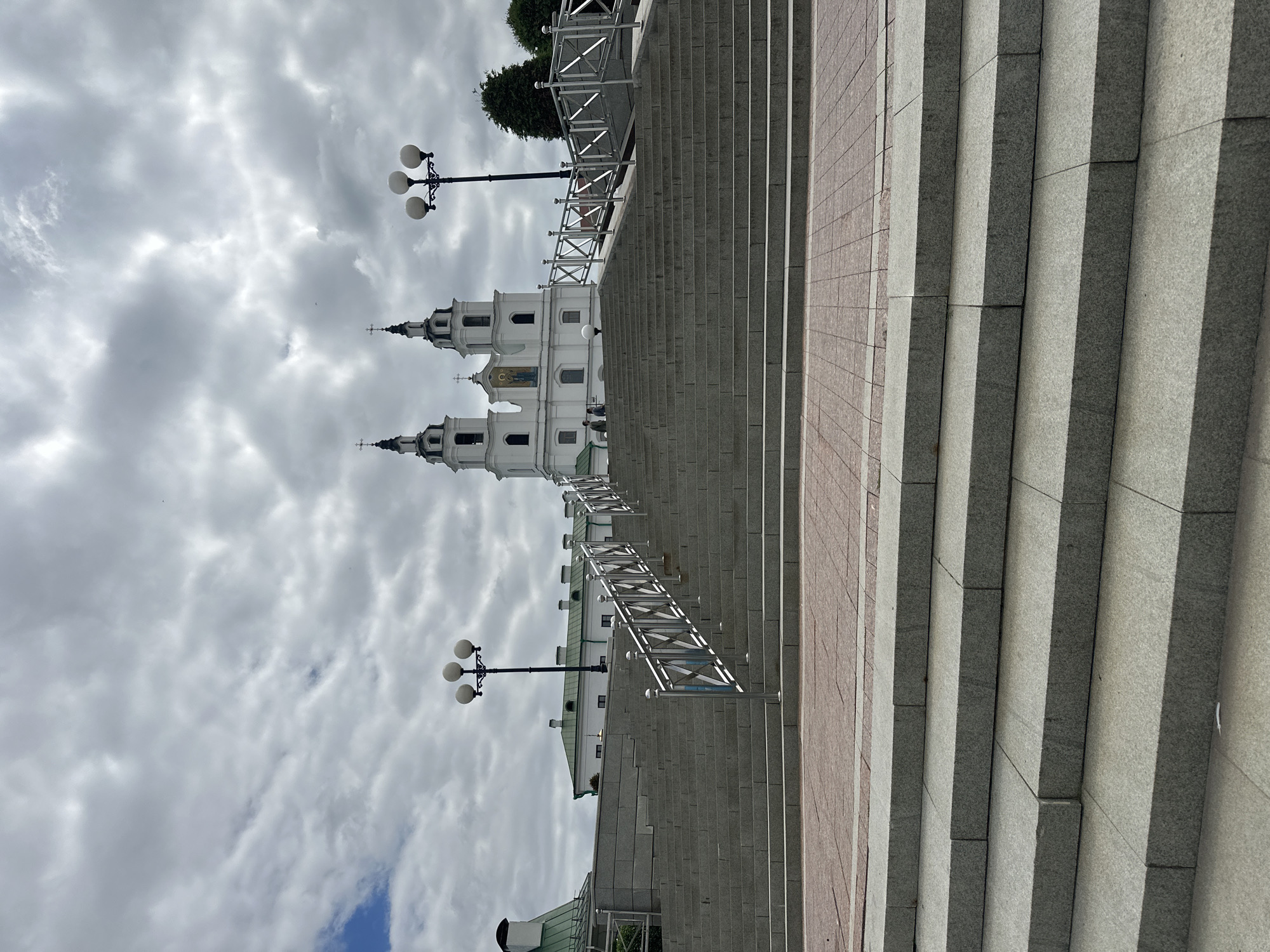 The image shows a wide, long staircase leading up to a large white building with two towers, located in the city of Minsk, Belarus. The building is likely a religious structure or a historical landmark, with a prominent cross on top of each tower and a religious fresco above the entrance. The staircase is made of gray stone slabs and features a metallic railing on both sides. At the top of the stairs, there is a walkway with lampposts. The sky is cloudy, and there are some trees visible in the background. Two individuals are seen walking on the upper part of the staircase, near the building.