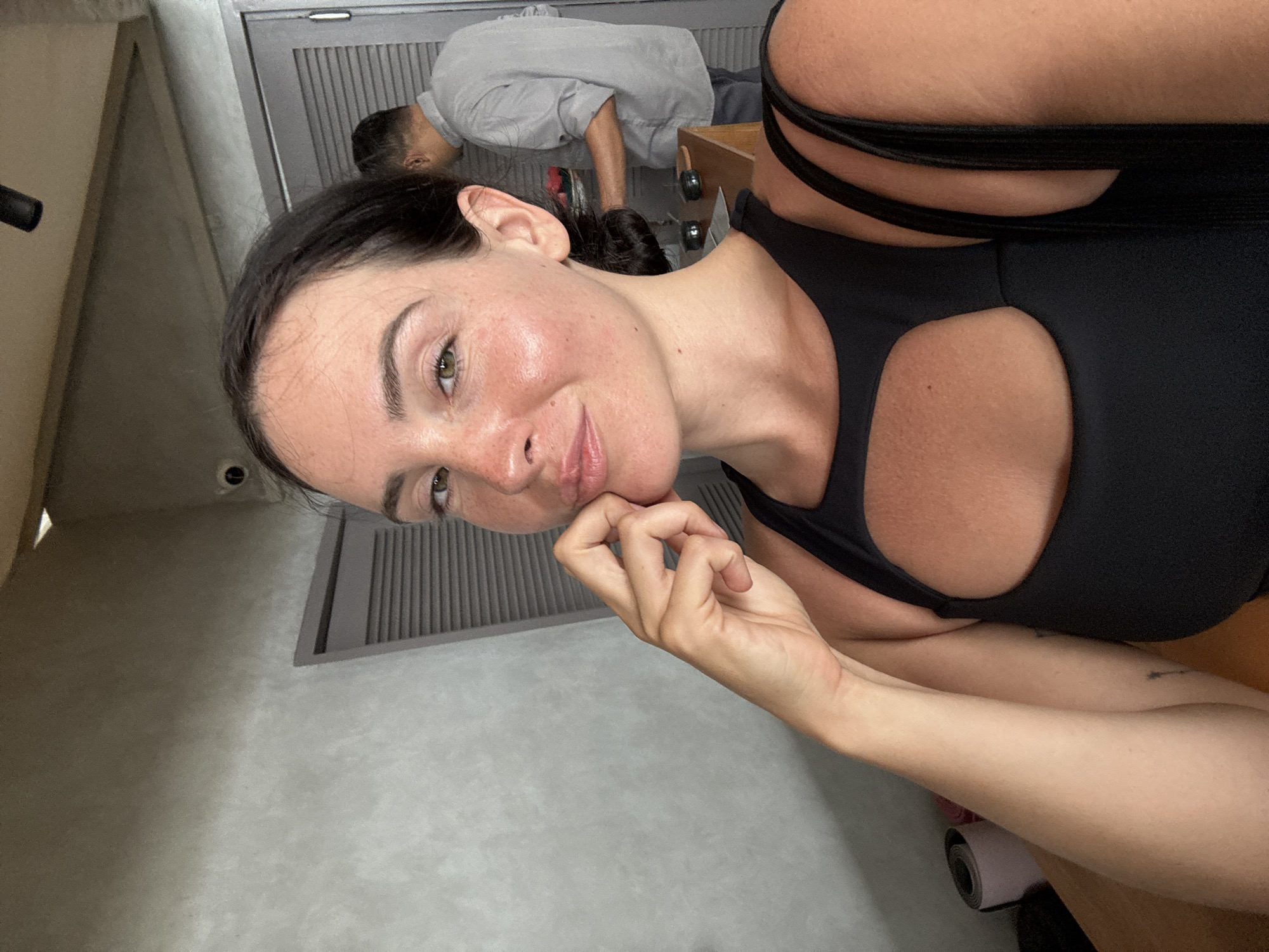 In North Kuta, Indonesia, a woman takes a selfie in a black workout top. She has brown hair pulled back and is resting her chin on her hand. Her skin is sun-kissed with freckles visible. In the background, a man in a gray shirt is visible, facing away from the camera, near a door with louvers. The environment appears to be indoors with a concrete wall and a wooden counter.