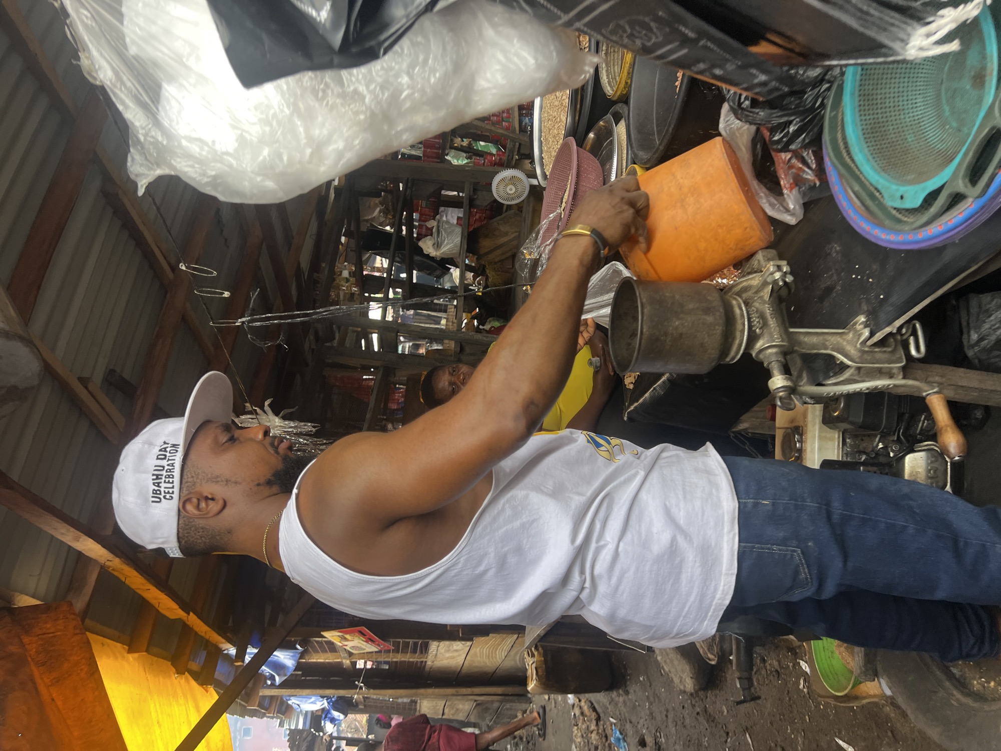 The image depicts an outdoor market scene in Nkpor, Nigeria, likely in a stall or shop with a corrugated metal roof supported by wooden beams. The ground appears to be dirt and debris. In the foreground, a man wearing a white tank top and blue jeans is visible from the chest down, facing away from the camera and to the right. He has a white baseball cap on with "UBAHU DAY CELEBRATION" printed on it. He is interacting with an orange bucket and a manual grinding machine, suggesting he is preparing or selling food items. His right arm is extended, and he is wearing a bracelet.
Behind him, partially obscured, is a woman wearing a yellow top, seated and looking towards the man. The stall is filled with various goods, including large plastic containers, baskets filled with what appear to be nuts or grains, and other unidentifiable items, some wrapped in plastic. A large white plastic bag is prominent on the right side of the frame, hanging from a wire. In the background, other market stalls and people can be seen, indicating a busy commercial environment. The overall impression is that of a functional, working market stall in a typical Nigerian setting.
