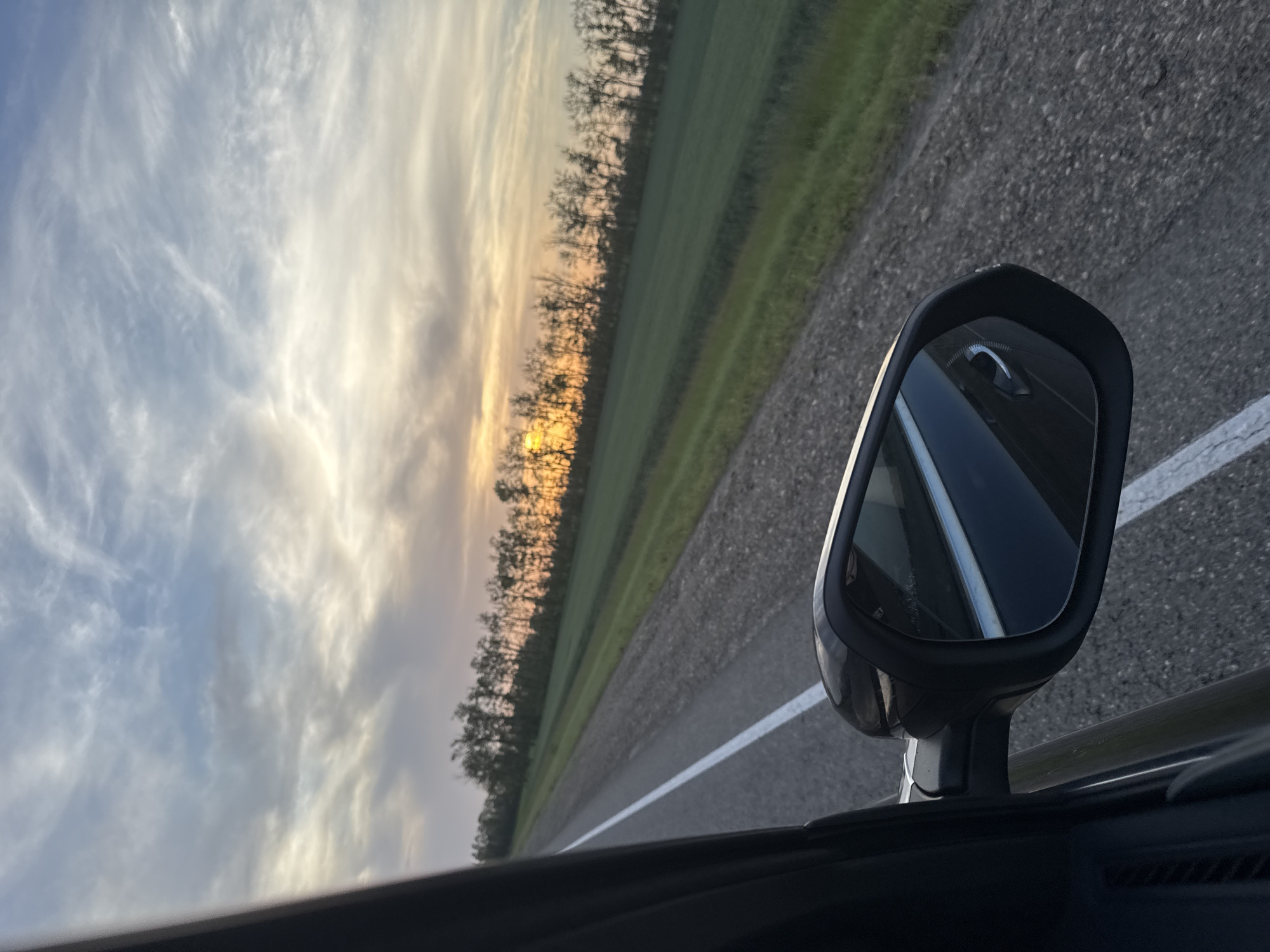 The image depicts a view from inside a moving vehicle, likely a car, driving on a rural road in Fastovetskaya, Russia. The road has a single white dividing line. Adjacent to the road is a strip of green land, bordered by a line of trees silhouetted against the sky at sunset/sunrise. The sky is visible with clouds. The car's side mirror is in the foreground, showing a reflection of the car's interior and the road behind. The asphalt appears to be in good condition. No people are visible in the frame.