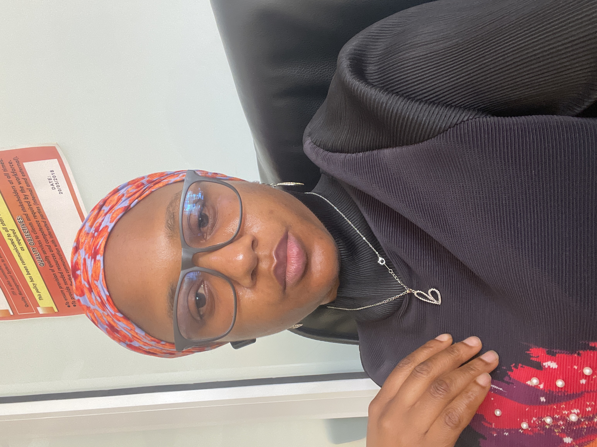 The image shows a woman wearing glasses and a colorful headscarf. She's wearing a black, textured turtleneck sweater with a silver heart-shaped pendant necklace. Her hand is resting on the sweater, which features red, orange, and purple patterns, accented with small pearls. The background includes a black leather chair and a document attached to a wall, which is titled "QUALITY OBJECTIVES." The date on the document is 20/05/2018. Based on the provided information, the image cannot be explicitly traced to Jos, Nigeria.