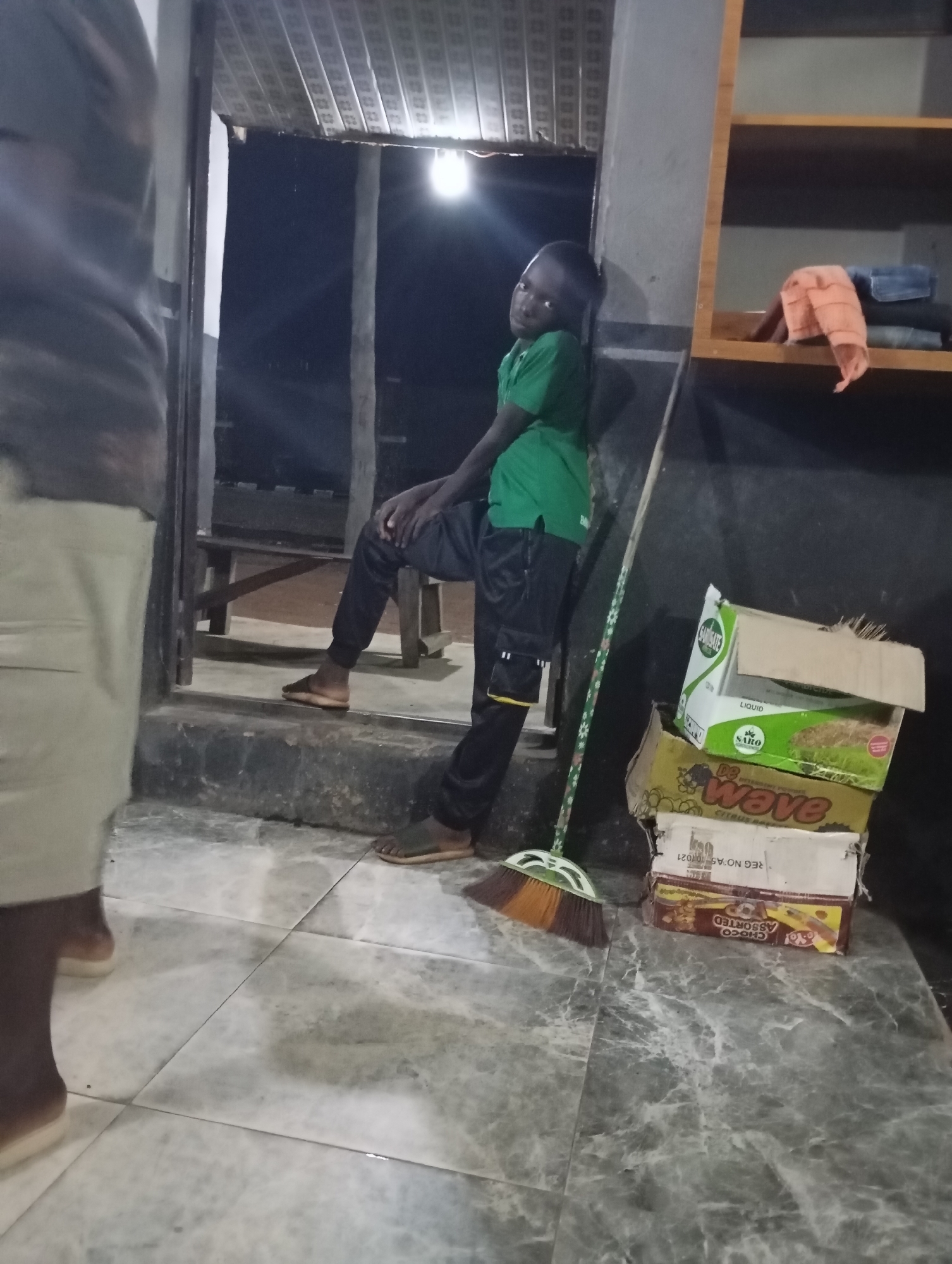 In Shendam, Nigeria, a young Black boy in a green polo shirt and black pants leans against a grey pillar. He is positioned in the foreground, to the left of a broom with a green and white patterned handle and brown and orange bristles. Beside him, a stack of cardboard boxes is visible. The top box is green and white with the text "SARGATE LIQUID" and "SARO". The box below it is yellow and features the word "wave" in red lettering. A third box, white and red, is partially obscured by the box above. The bottom box, also partially visible, is brown and has an image of a banana and text that includes "CHOCO ASSORTED". In the background, a light source illuminates the scene, and a shelf unit with folded clothes, including an orange cloth and blue denim, is present. The floor is tiled with a grey and white marbled pattern. Another person, only their legs and feet are visible, stands to the left, wearing light-colored trousers and flip-flops. A wooden bench is also present in the background.