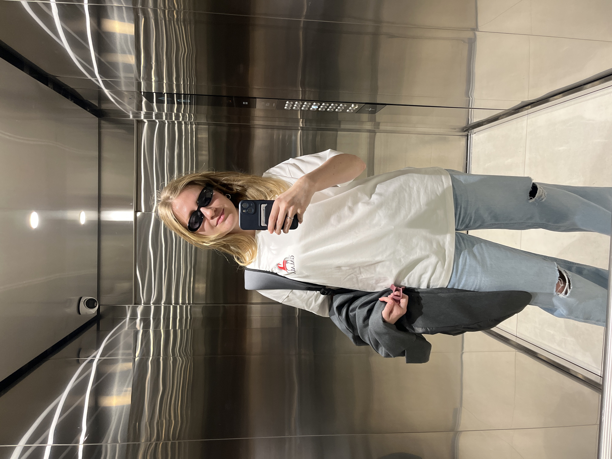 A young blonde woman with sunglasses takes a selfie in an elevator. She wears a white t-shirt and ripped blue jeans, holding a black smartphone. A grey jacket hangs from her shoulder. The elevator has stainless steel walls, a row of illuminated buttons, and a security camera in the upper left. The floor is light-colored tile. The image provides no information to identify its location in Belgrade, Serbia.