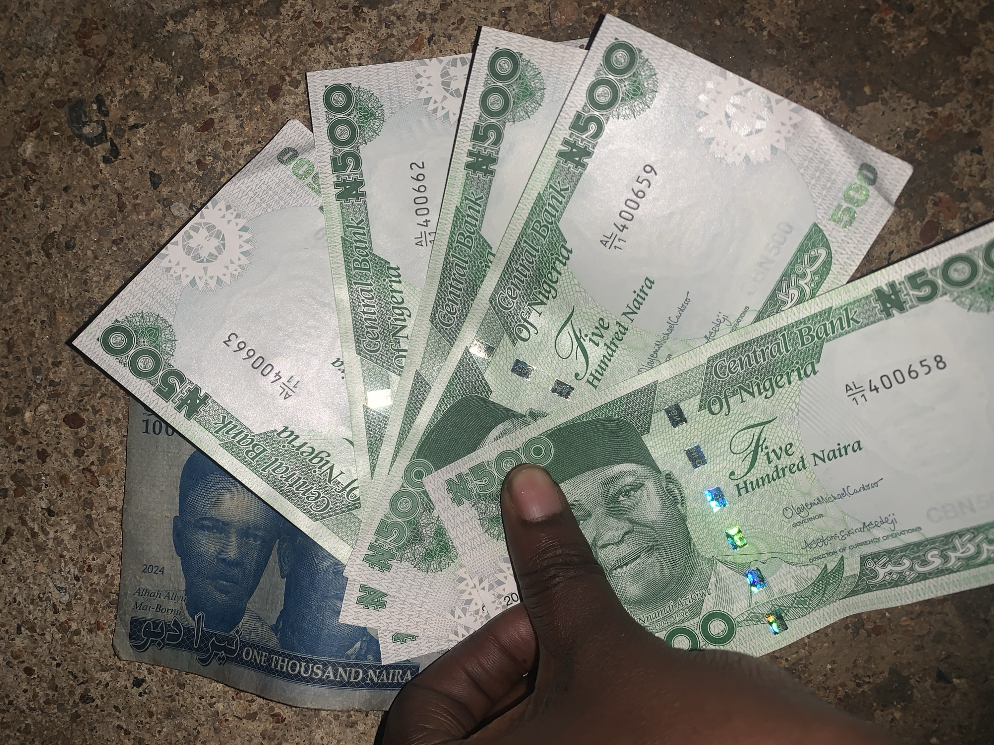The image shows a hand holding several Nigerian Naira banknotes on a textured surface. There are five 500 Naira notes and one 1000 Naira note partially visible. The 500 Naira notes are predominantly white with green accents and feature the portrait of a Nigerian leader. The 1000 Naira note is blue and features two portraits. The serial numbers on the 500 Naira notes visible are AL/11 400662, AL/11 400659 and AL/11 400658. The serial number on the 1000 Naira note is AL/11 400663.
There is no evidence that supports the location of New Stadium in Nigeria.