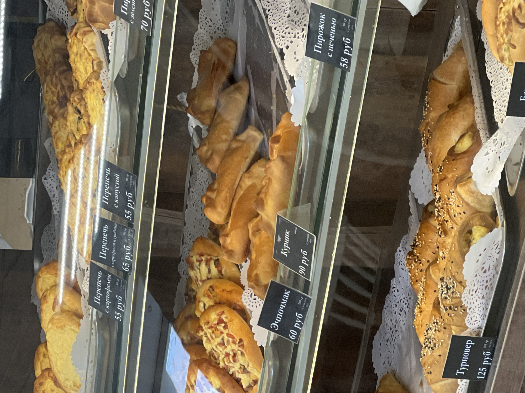The image depicts a display case of various baked goods with price tags, likely in a bakery or cafe. The price tags are in Russian rubles and some of them read: "Perepech" with different fillings (potato, potato and mushrooms, cabbage), "Echpochmak", "Kurnik", "Turnover", "Pirozhok" with liver. The baked goods are arranged on lacy paper doilies.